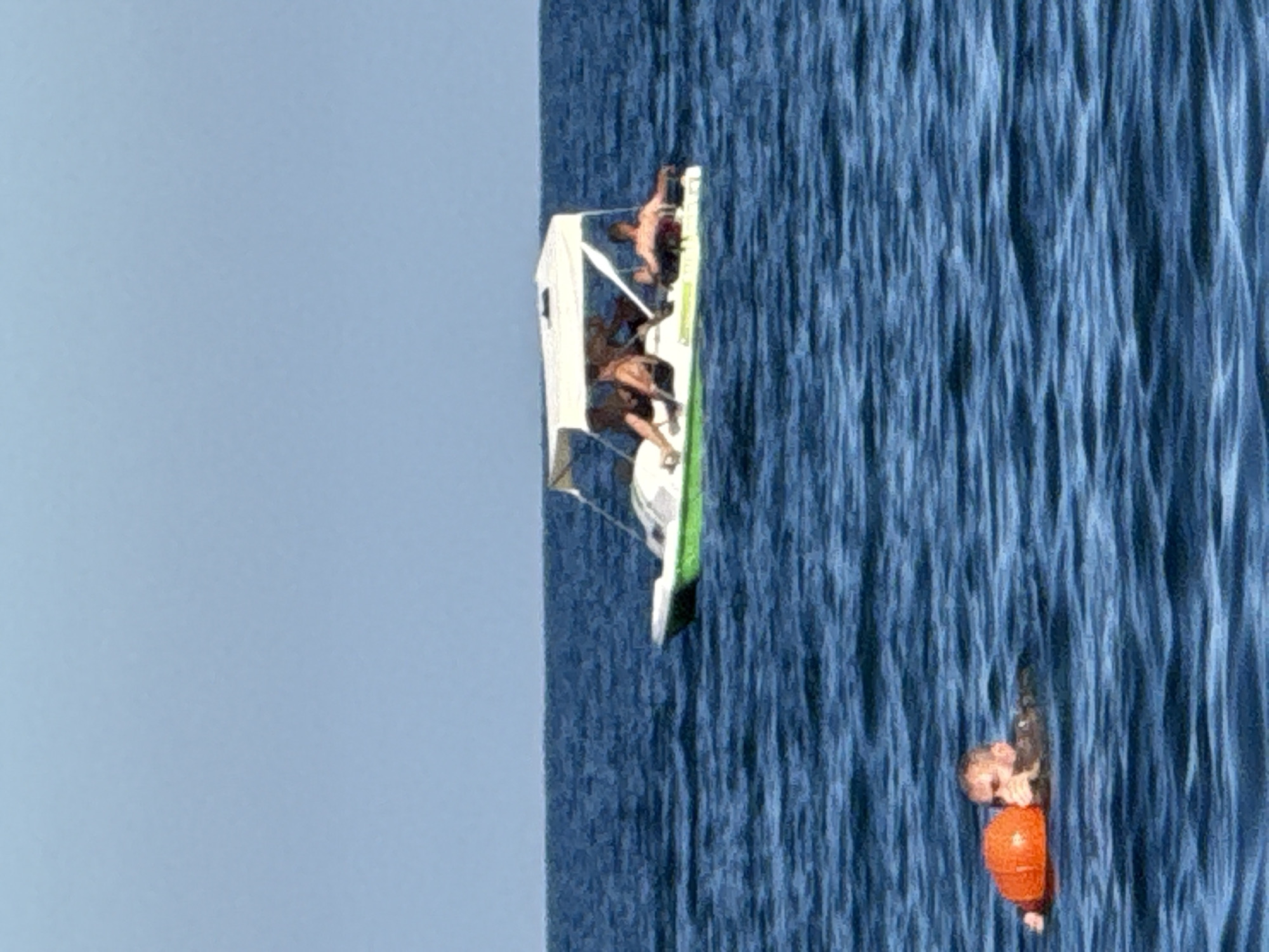 In the waters off the coast of Partenit, Ukraine, a man is seen swimming in the sea, holding onto an orange flotation device. Further out in the water, a pedal boat with a canopy is carrying several individuals, some of whom appear to be sunbathing. The sea is a deep blue with visible ripples, and the sky above is a clear, light blue.
