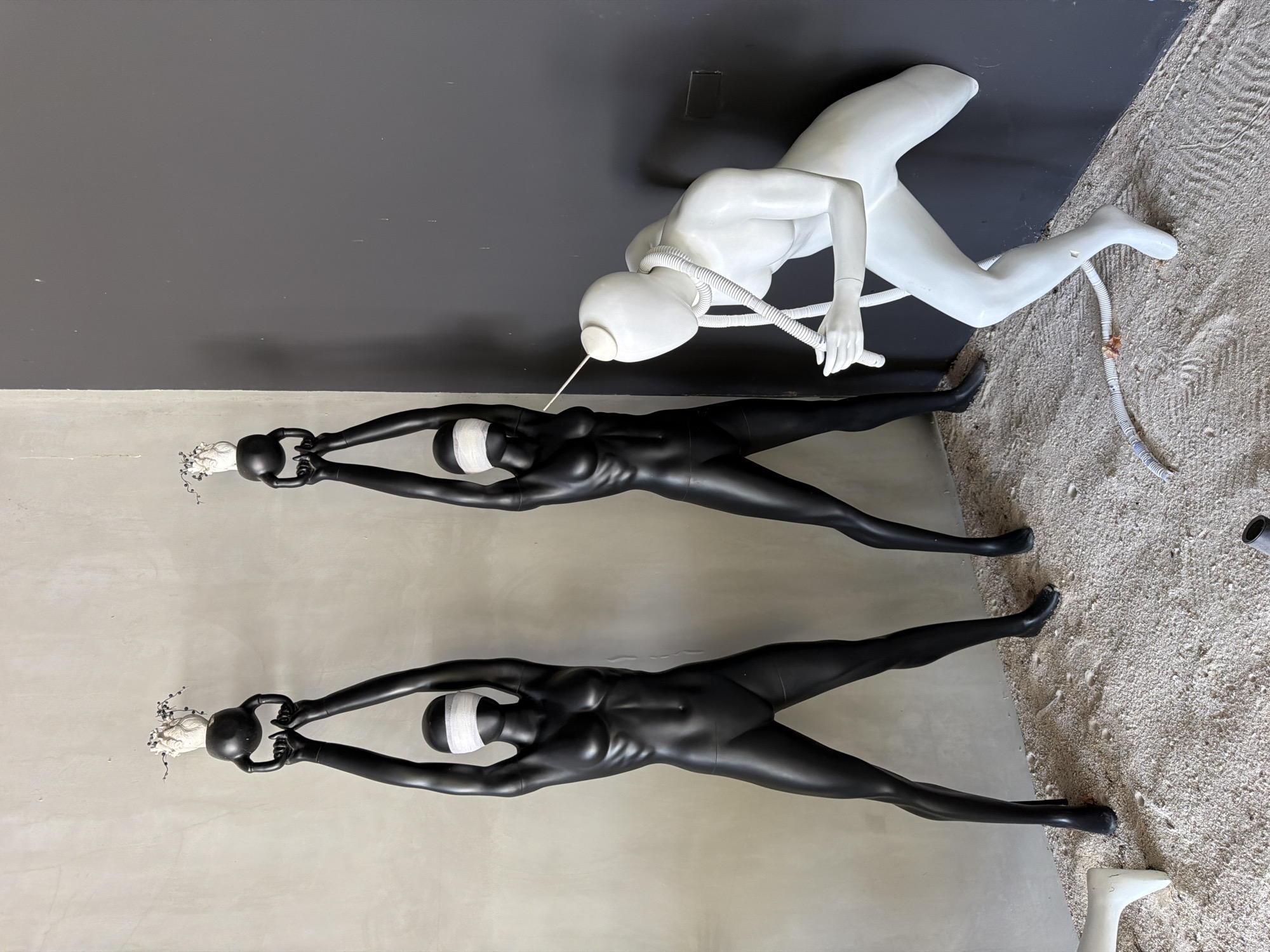 The image depicts an art installation in Ko Samui District, Thailand, featuring three mannequins on a sandy floor in front of a textured wall and a dark gray wall. Two identical black female mannequins stand upright, each blindfolded with white fabric, holding a kettlebell above their heads. The kettlebells are topped with abstract white sculptures resembling hearts with small branches and berries. To the right, a white male mannequin is in a crouched position, facing the black mannequins. A thick white tube or hose is wrapped around the white mannequin's neck and is also connected to a thin white cord that extends towards the black mannequin on the left. The white mannequin's hands are positioned as if manipulating the tube or cord. The environment suggests an indoor space with a rough, unfinished aesthetic, possibly an art gallery or studio.