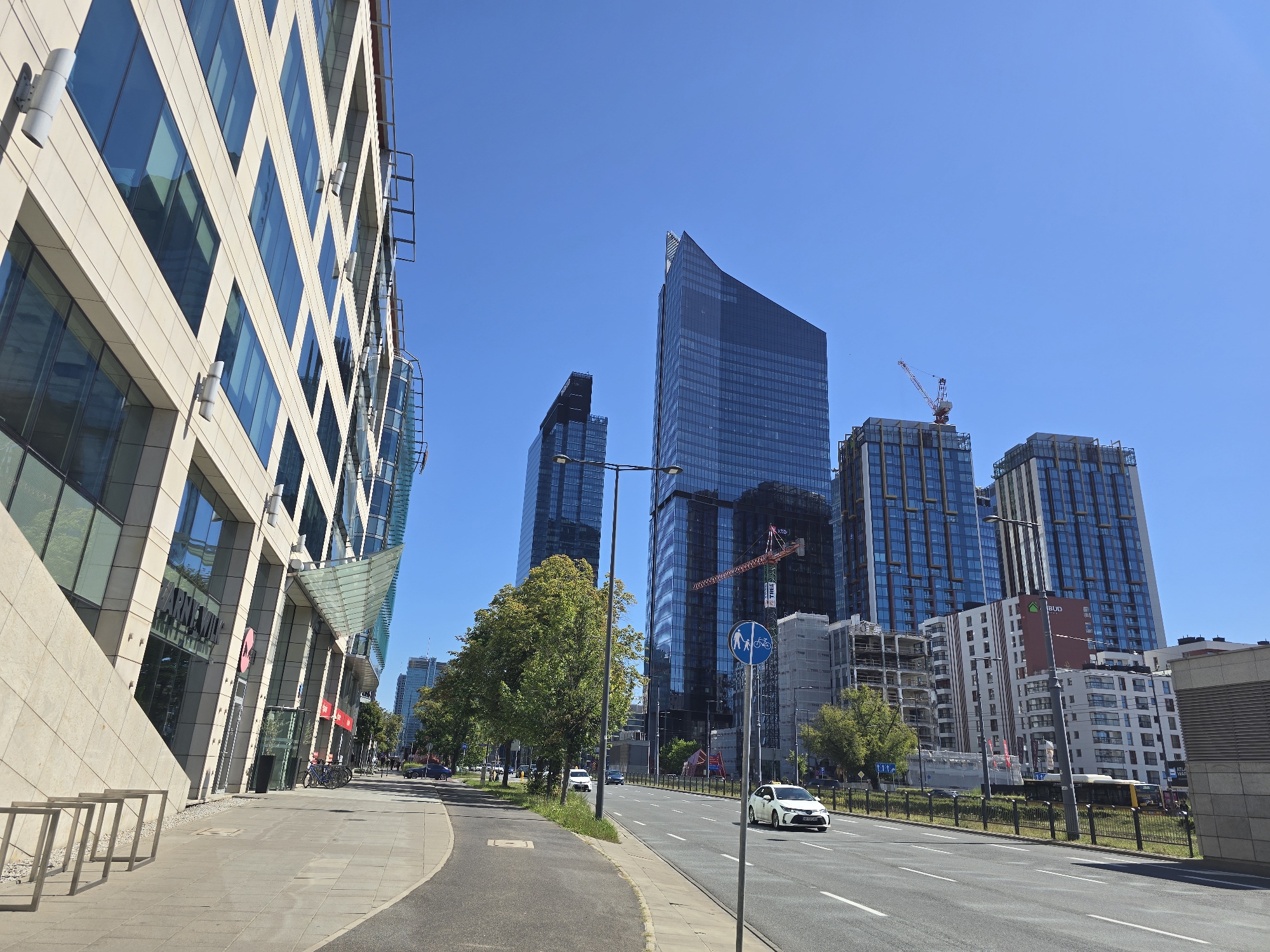 This is a daytime, eye-level, medium shot of a street scene in Warsaw, Poland, likely in the city center or a modern business district. The image captures a wide, multi-lane road with a clear blue sky overhead.
In the foreground, a light-colored, modern building with a beige stone facade and numerous glass windows dominates the left side of the frame. The building has a dynamic, angled design with glass awnings over some entrances. A paved sidewalk runs alongside the building, featuring bicycle racks.
To the right of the sidewalk, the road stretches into the distance. Several cars are visible, including a white sedan and a taxi. Trees line the side of the road, with lush green foliage.
The background is characterized by a cluster of tall, contemporary skyscrapers with dark glass facades. Some of these buildings appear to be newly constructed or still under construction, with scaffolding visible. A red construction crane is prominent among the buildings. Traffic signs, including one indicating pedestrian and bicycle paths, are also present.
The overall impression is of a modern, urban environment on a sunny day.