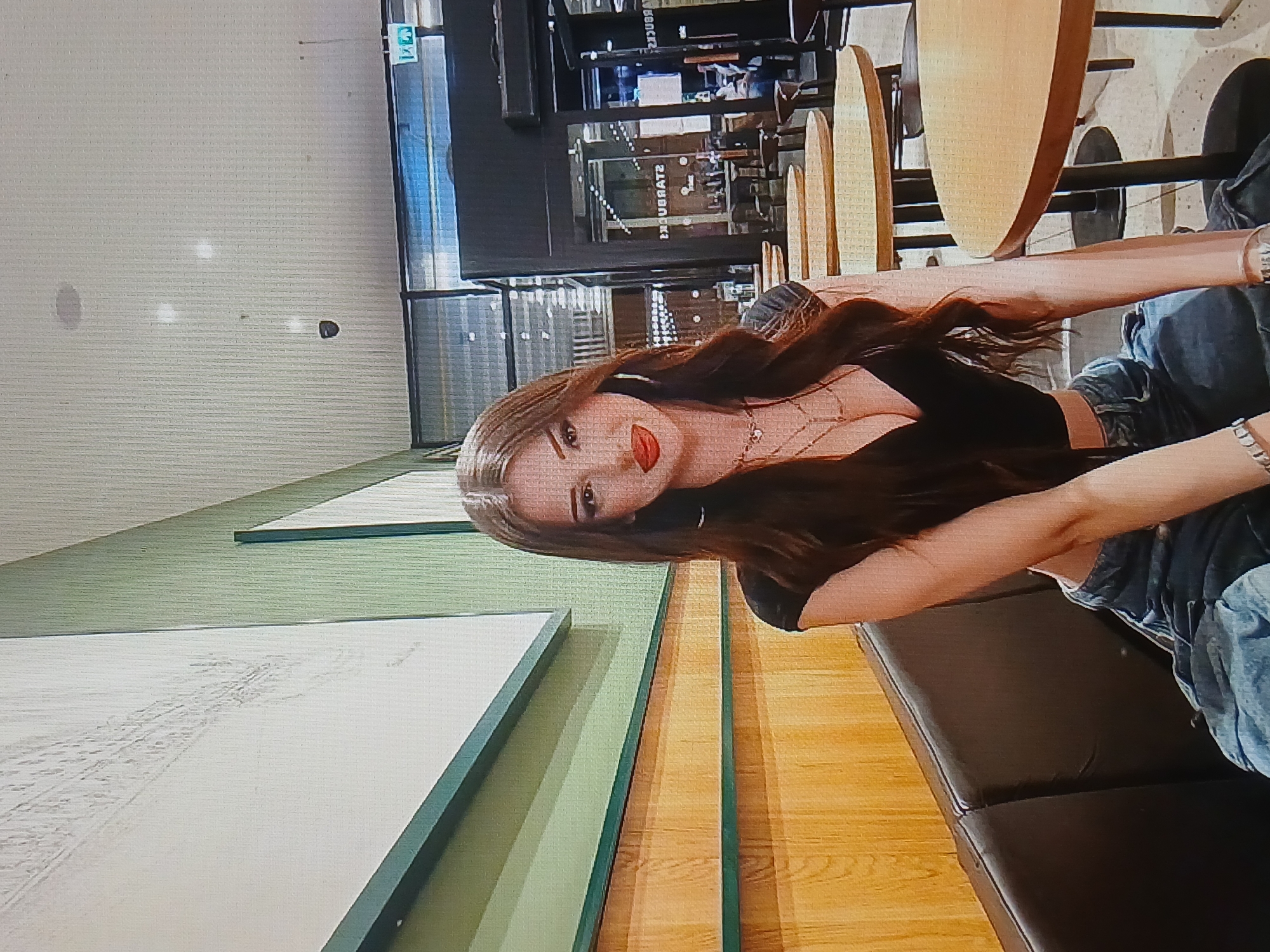A young woman with long, dark hair and red lipstick is seated on a dark leather bench in what appears to be a Starbucks. She is wearing a black, low-cut top and distressed denim jeans. The background shows a typical Starbucks interior with wooden tables and chairs, and large windows. The woman is looking directly at the camera with a slight smile. The location is identified as Dnipro, Ukraine.