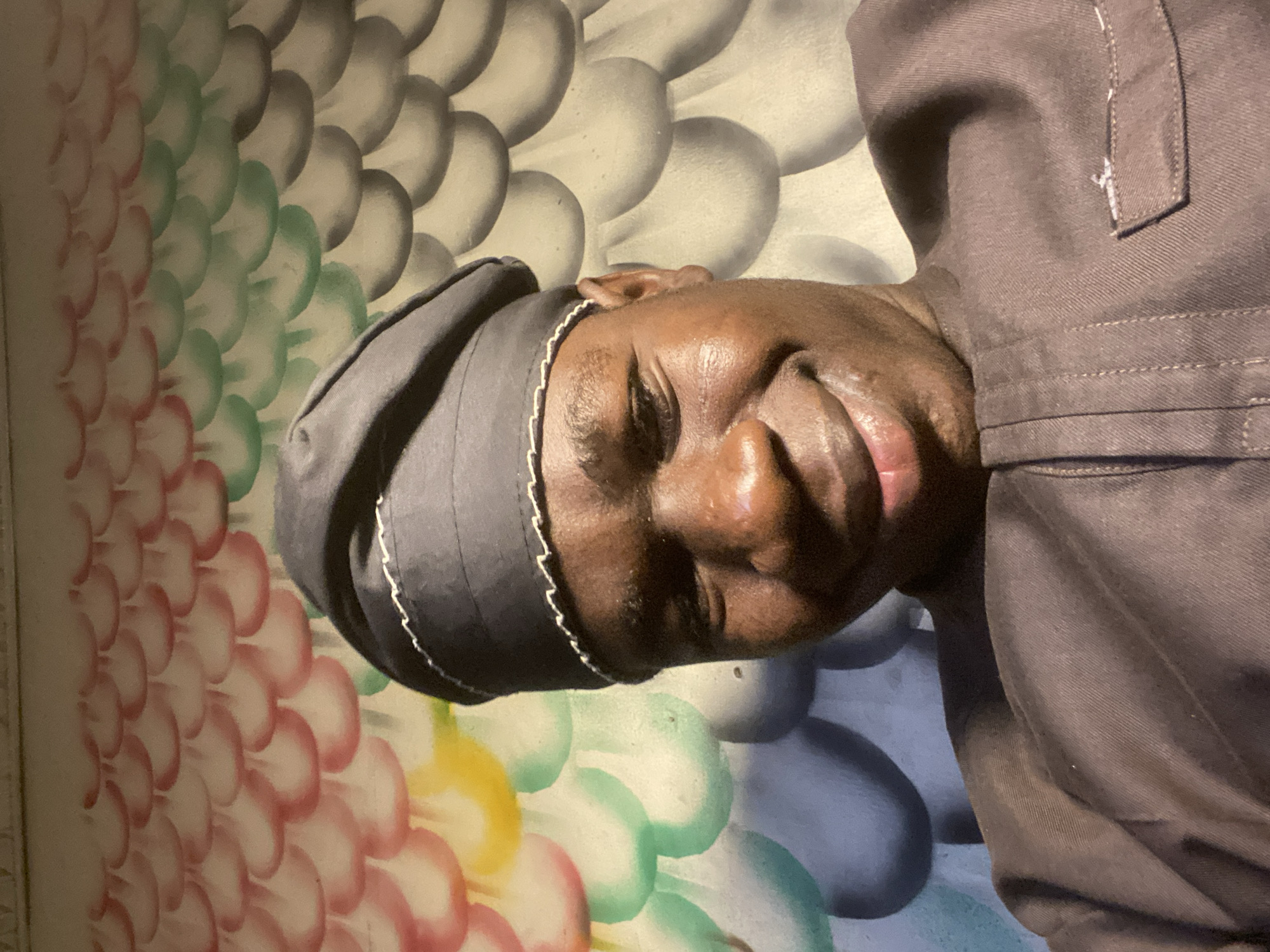The media file presents a close-up portrait of an individual with dark skin, resting or sleeping, with the image oriented sideways. The person's head is on the left side of the frame, facing right, with eyes closed and a relaxed expression on their face, indicating a state of peace or repose. They are wearing a dark cap or head covering with a light-colored, possibly white, trim detail, and a dark brown garment, likely a shirt or jacket, visible on their upper body.
The background features a distinctive pattern composed of overlapping, scale-like or petal-like shapes arranged in rows. This pattern displays a gradient of colors, transitioning from reddish-pink on the far left, through shades of green in the center, to greyish-white on the right side behind the person's head and shoulder. A small section with a yellow tint is also discernible towards the bottom left of the pattern. The lighting in the scene is soft and appears somewhat dim. There is no visible text in the image. The setting suggests an indoor environment against a decorative wall or backdrop.