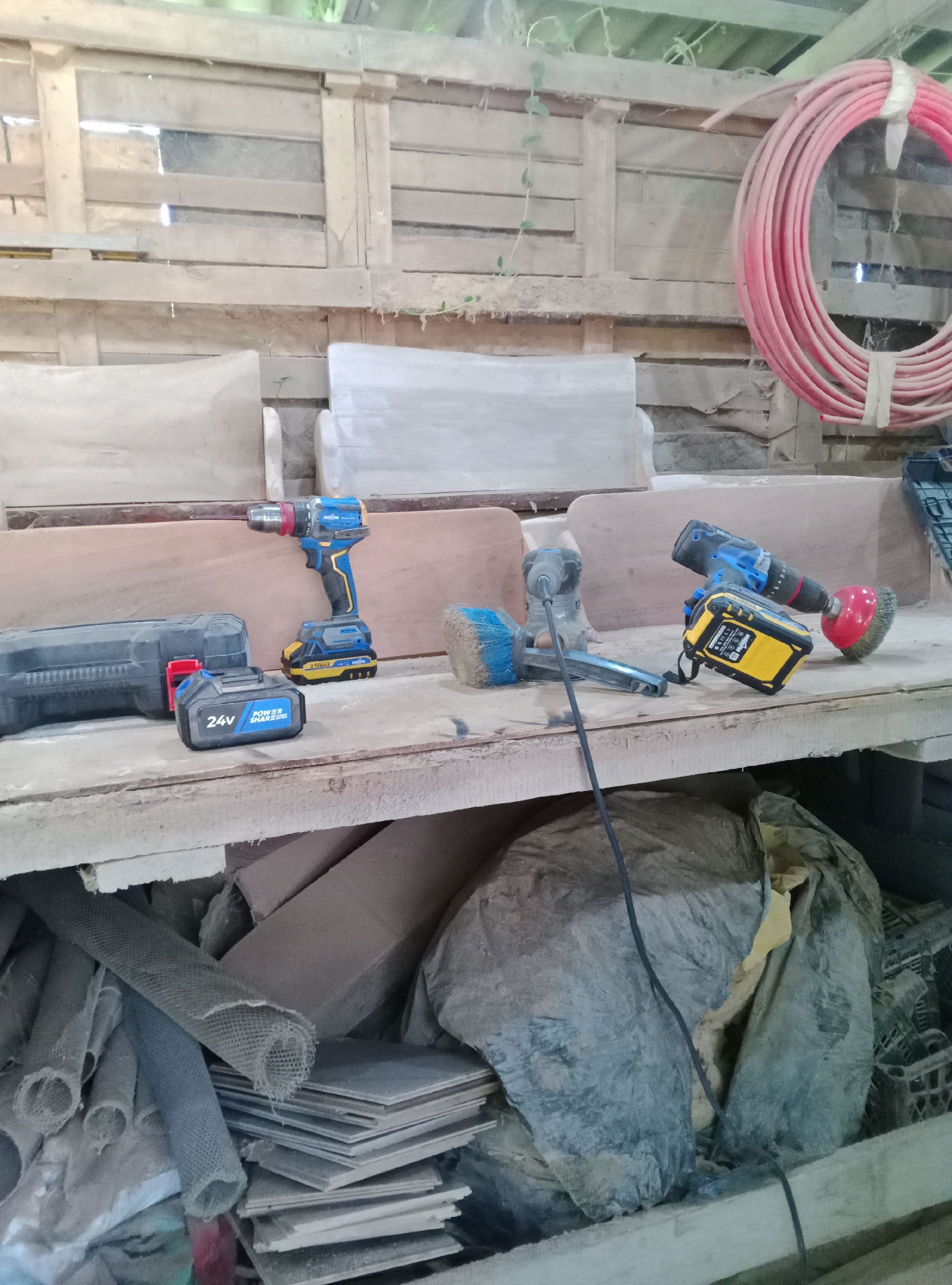 This image depicts a workbench in what appears to be a workshop or storage area. The workbench is made of weathered wood and is cluttered with various tools and materials. In the foreground, a blue and yellow power drill with a battery is visible. To its left, a black case and a detached 24V battery are present. Further to the right, another power tool with a brush attachment and a yellow battery is in view, along with a red-capped wire brush accessory.
Behind the workbench, wooden pallets form the background. A coiled red hose is hanging on the wall to the right. The environment appears dusty and somewhat disorganized, with piles of what look like cardboard, mesh material, and large plastic bags on the lower shelves and floor. The overall impression is of a working space that is actively used. The location is stated to be in Pshap, Abkhazia, suggesting this is an outbuilding or workshop in that region. There are no people visible in the image.