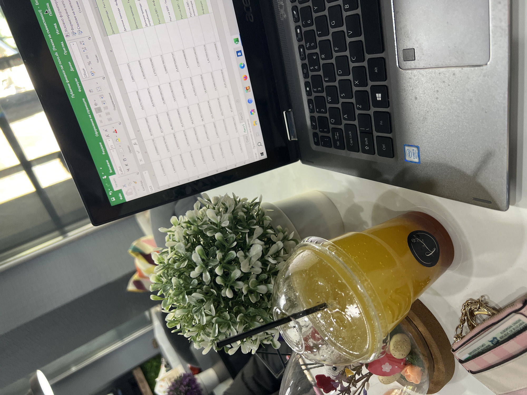 This angled overhead shot captures a desktop setup in what appears to be an indoor setting, possibly a cafe or office, during daylight hours.
The central subject is a silver Acer laptop, an Intel Core i5 8th Gen model, open to a Microsoft Excel spreadsheet. The spreadsheet displays columns with numerical data and headers in Bulgarian, including "Данъчни приходи" (Tax revenues), "Ръст на БВП" (GDP growth), and "Инфлация" (Inflation). The Excel interface also shows Bulgarian menu options such as "Файл" (File), "Начало" (Home), and "Данни" (Data). The laptop's keyboard features both Latin (QWERTY) and Cyrillic characters.
To the right of the laptop is a clear plastic cup filled with a vibrant yellow/orange beverage and ice, accompanied by a black straw. A black circular sticker with a stylized 'L' logo is visible on the cup. Nearby, a small artificial plant with green and white foliage sits in a white pot. Further to the right, there's a decorative cloche containing miniature, colorful items resembling macarons or cookies and tiny artificial flowers. A small pink and white striped wallet or cardholder with a gold chain is placed beside the cloche. Text on a card inside the wallet clearly reads "EUROPEAN UNION" and "REPUBLIC OF BULGARIA," aligning with the provided location context of Plovdiv, Bulgaria.
The arrangement suggests an individual is engaged in work or study, taking a break with a drink in a casual yet organized environment.