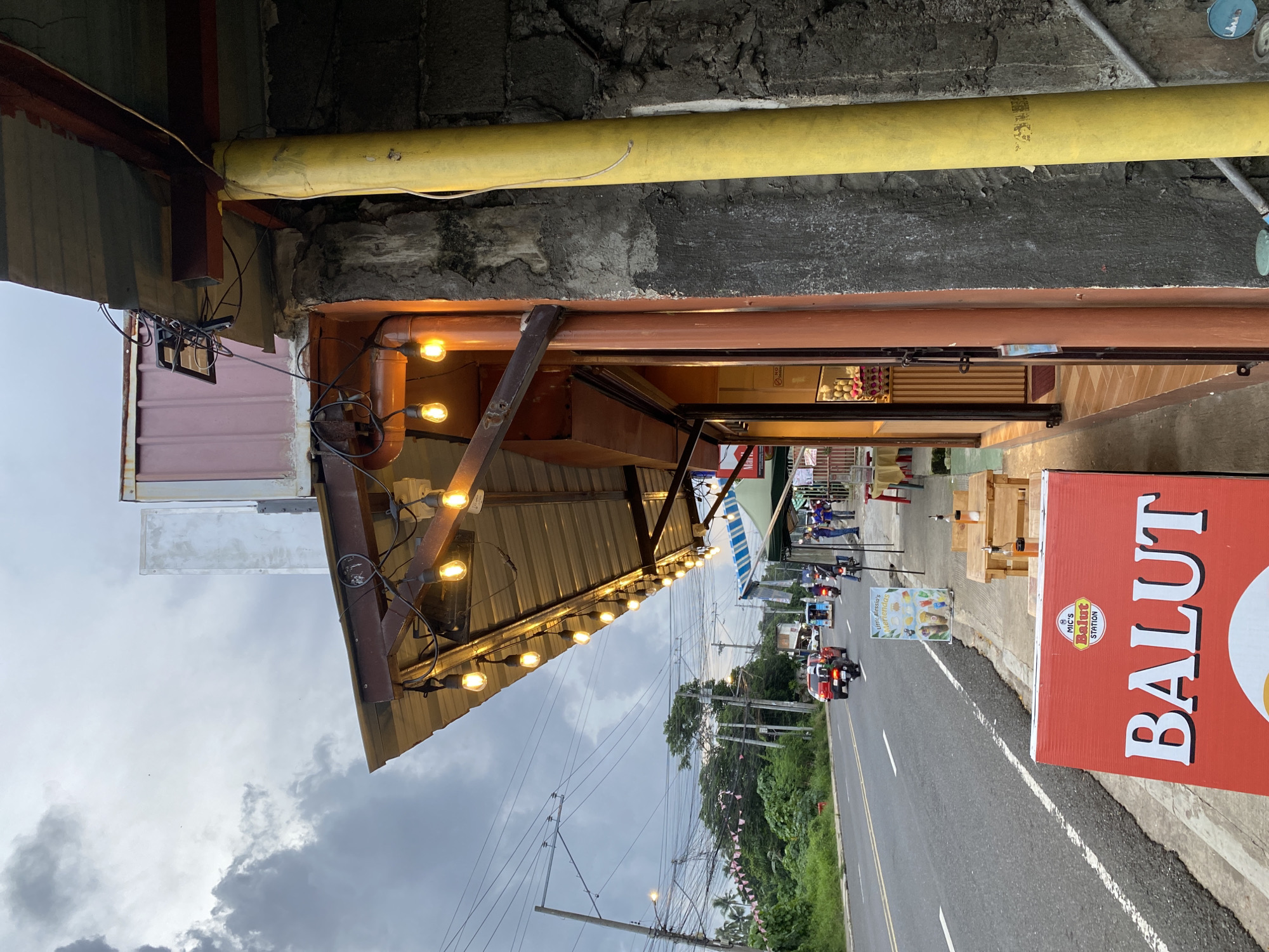 This is an eye-level, medium shot of a roadside eatery in Davao City, Philippines, under a cloudy sky. The foreground is dominated by a bright red sign advertising "BALUT" from "MIC'S BALUT STATION". To the left of the sign is a two-tiered wooden table with salt and pepper shakers. Behind the table, the eatery's facade is visible, featuring orange-painted poles and a corrugated metal awning adorned with a string of illuminated bulbs. A glass display case inside showcases fruits.
The middle ground shows a busy asphalt road with white lane markings, lined with trees and utility poles. Several vehicles, including motorcycles and cars, are traveling on the road. A white and blue striped awning extends from a stall on the right side of the road.
The background features a dense canopy of trees and more utility poles, suggesting a tropical urban environment. The overall lighting is dim, indicative of overcast weather.