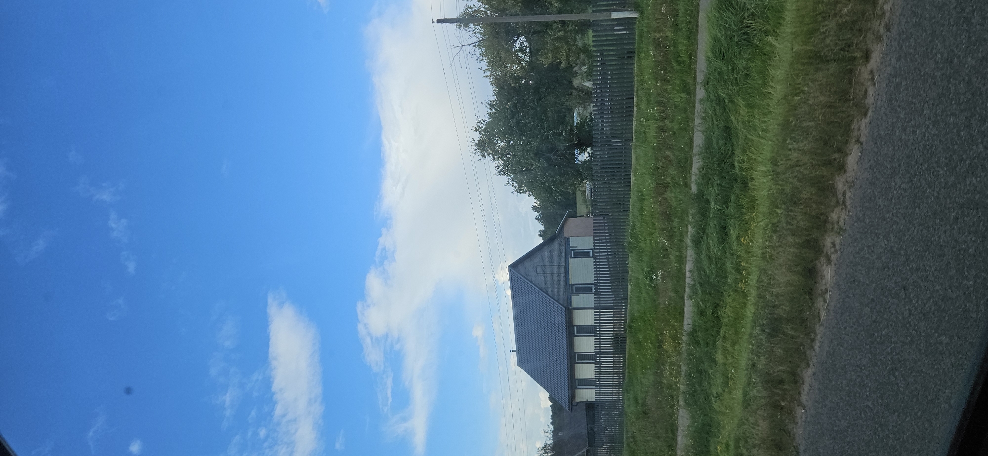 A single-story house with a dark, pitched roof and light-colored siding is viewed from the side, behind a wooden fence. The house has several windows with dark frames. There is a large tree to the right of the house, and a telephone pole with wires. The house is surrounded by green grass, and a paved road is visible in the foreground. The sky is blue with scattered white clouds. The scene appears to be a rural setting in the Варонаўскі сельскі Савет, Belarus.