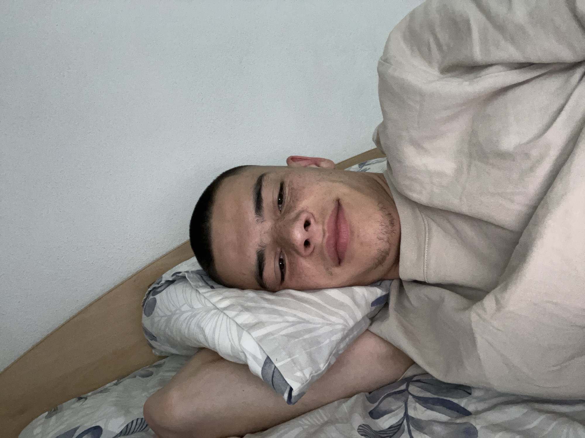 A young adult male is lying in bed. He is resting his head on a pillow patterned with gray and white leaves. The bedsheets also feature a leaf pattern. He is partially covered by a light-colored blanket or sheet. The room has plain white walls, and a wooden headboard is visible behind the pillow. The location cannot be determined to be in Karavelovo, Bulgaria.