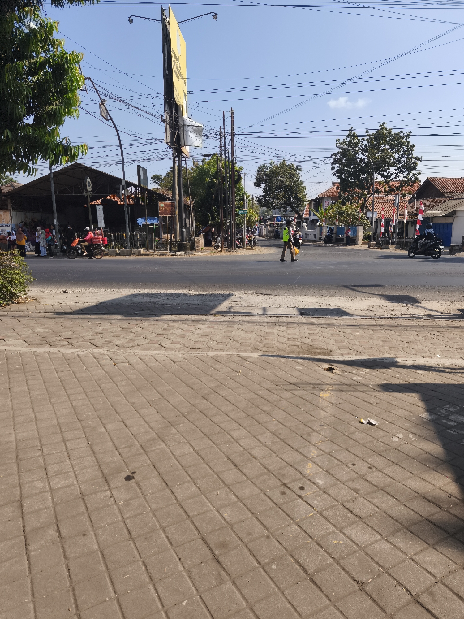 The image depicts a street scene in Kutoarjo, Indonesia. A paved sidewalk runs in the foreground, with a street beyond it. Several people are visible on the side of the street, some standing and some on motorcycles. A man in a bright yellow vest is walking across the street. Buildings line the opposite side of the street, with trees and utility poles scattered throughout the scene. The sky is clear and blue, indicating a sunny day. Indonesian flags are visible, suggesting a national holiday or event. A large billboard stands prominently on the left side of the image. The overall impression is that of a busy, everyday street in a town or city.
