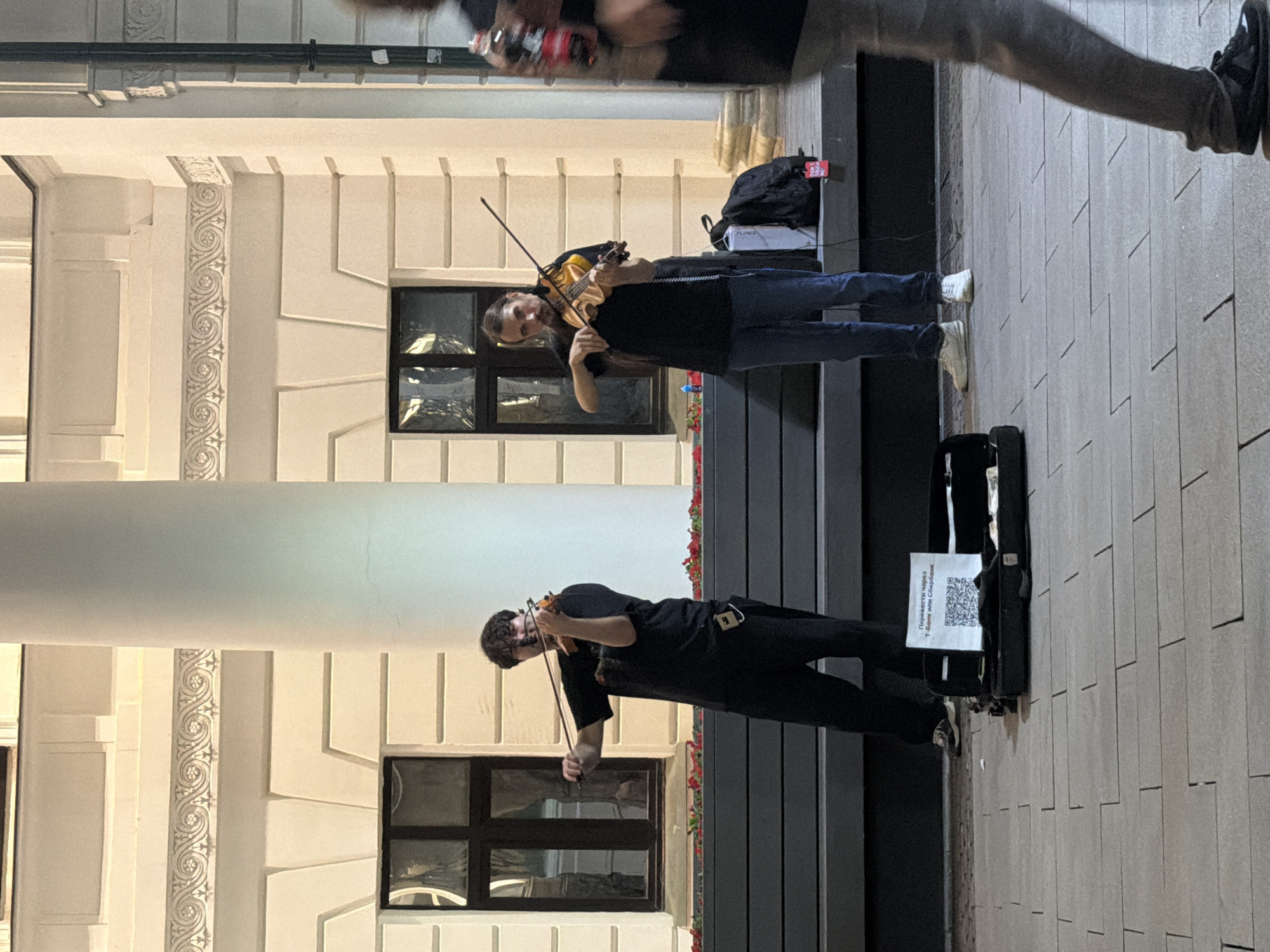 The image depicts an outdoor street scene with two men playing violins. They are standing in front of a white building with classical architectural elements, including columns and decorative molding. The ground is paved with grey rectangular tiles. A black violin case lies open on the ground in front of the musicians, displaying a QR code and some Cyrillic text, suggesting a method for receiving donations. One musician, on the left, wears a black t-shirt and black pants, while the other, on the right, wears a black t-shirt and blue jeans. Both are actively playing their instruments. In the foreground, the legs of a person walking by are visible, partially obscuring the scene. The lighting suggests it might be evening or late afternoon. The building's facade indicates a potentially historical or significant urban area. The presence of a QR code for donations is a common feature of street performers. The scene is likely located in a public space in Nizhny Novgorod, Russia, given the Cyrillic text on the donation sign.