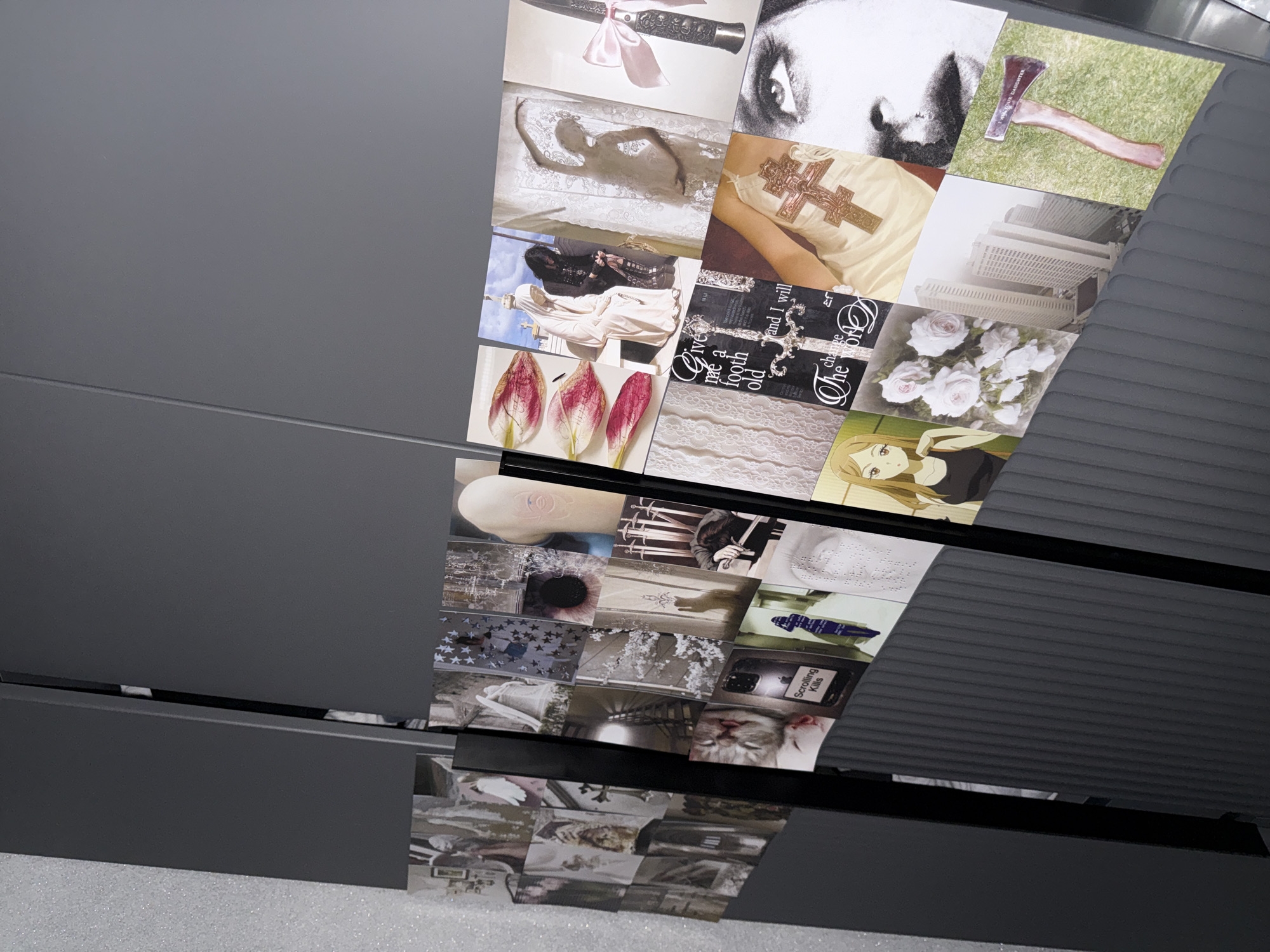 A multi-level display unit, possibly a set of pull-out drawers or shelves, showcases a diverse collage of images. The unit itself is a dark gray or black, with smooth, flat surfaces and ribbed sections on the right side. The images, varying in size and orientation, are affixed to the internal surfaces of what appear to be opened compartments.
The collage features a wide array of subjects and objects. Visible images include:
* A knife with a pink bow.
* A figure resembling a mummified body on a white lace fabric.
* A close-up of a human eye in black and white.
* An axe lying on green grass, with the text "MOTHER DAUGHTER" visible on its handle.
* A person wearing a light-colored top with a large, ornate cross.
* An old computer monitor.
* A cluster of white roses.
* Three red tulip petals.
* An image with a large, ornate key or cross and partially visible text: "Give me a foote old and I will change The world."
* White lace fabric.
* An anime-style illustration of a girl.
* A pale arm or leg with "CHANEL" inscribed or marked on it.
* Several swords.
* A person with dark hair, head down.
* A white surface with braille dots.
* A phone screen displaying the text "Scrolling Kills."
* A sleeping kitten.
* Various other abstract patterns, textures, and ambiguous forms.
The overall impression is that of a curated collection, possibly a mood board or a personal art installation, without a singular activity taking place. The lighting suggests an indoor setting.
