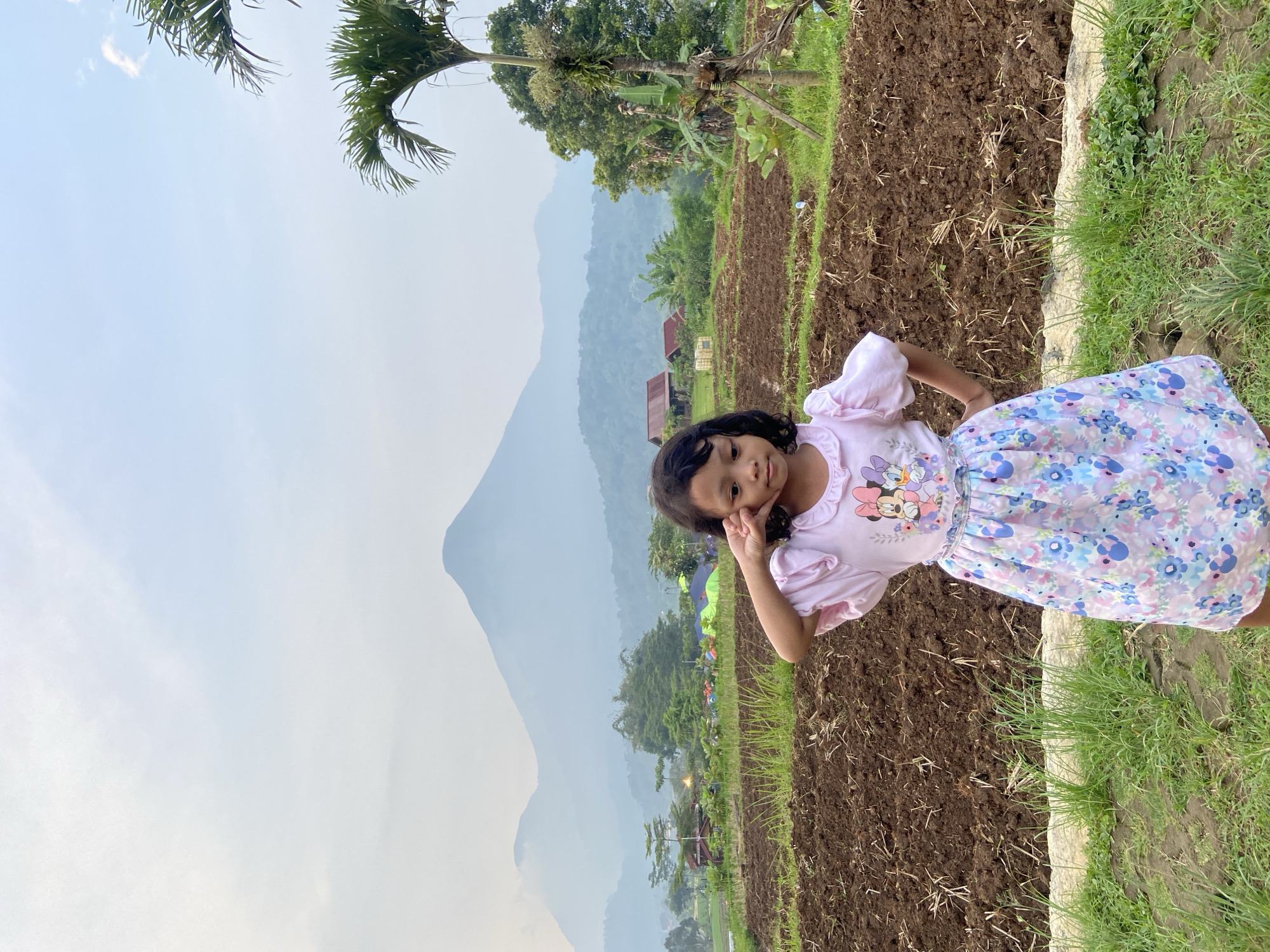 The image depicts a young girl standing in a field. She has dark hair and is wearing a light pink shirt with a floral skirt. She is posed with her hand near her face.

Behind her, the field is plowed into rows of dark soil. Beyond the field, there is a mountain range shrouded in mist. The sky is light blue. There are trees visible in the mid-ground. Structures suggesting human activity are present.

Based on the location tags, this scene likely takes place near Selotapak, Indonesia. The landscape includes cultivated land and natural mountainous terrain, with clear human development nearby.