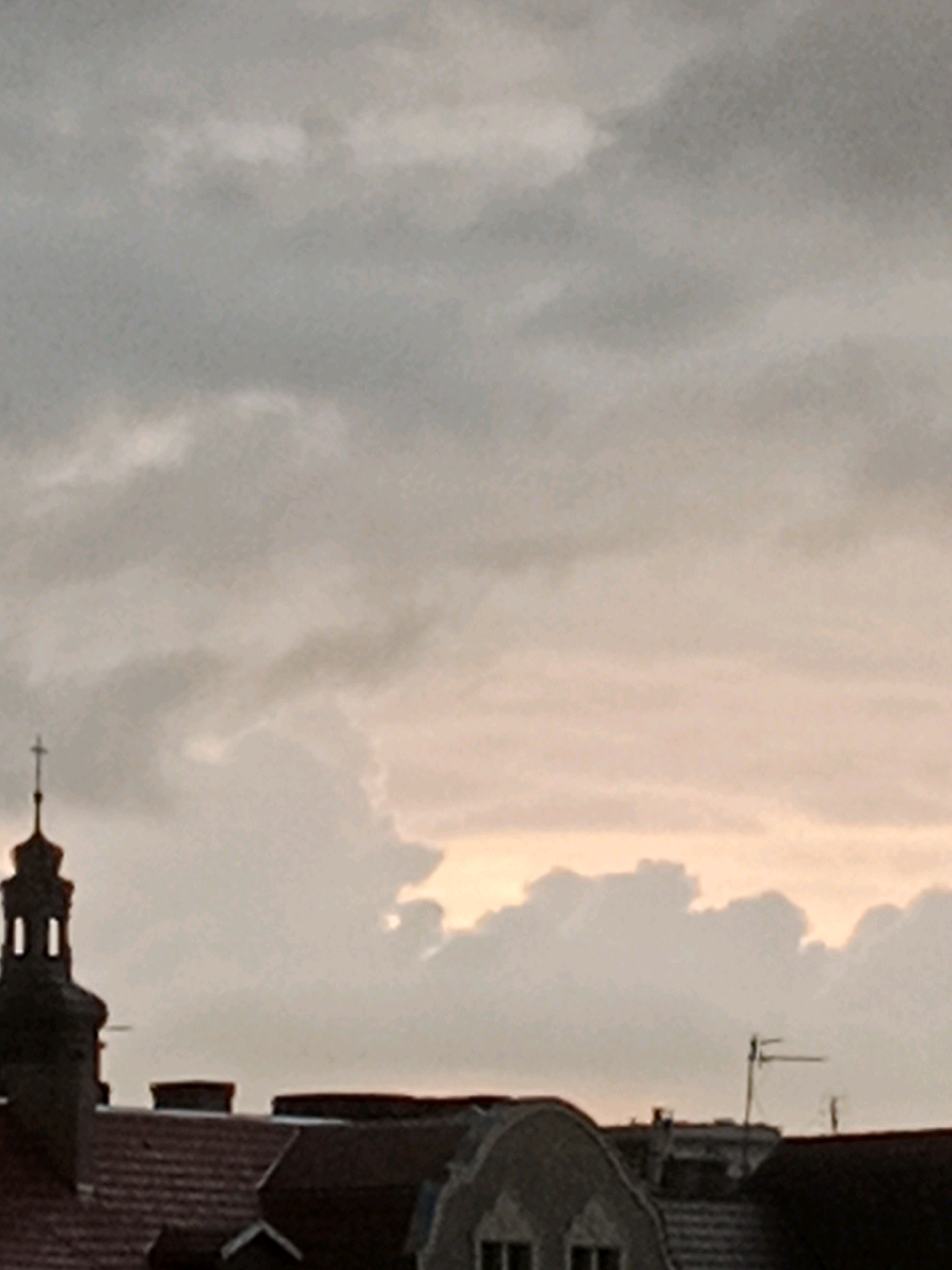The image captures a view of rooftops and a church spire against a cloudy sky, suggesting an overcast day with potentially breaking clouds towards the horizon. The scene appears to be an urban environment, likely in Bydgoszcz, Poland, given the architectural style of the church and the rooftops.
The prominent feature on the left is a dark-colored church tower, characterized by a pointed steeple topped with a cross. The tower has a few windows visible, suggesting internal illumination or empty openings. Below the spire, the tower has a tiered structure.
In the foreground, various rooftops are visible. They are predominantly tiled in shades of reddish-brown. Several chimneys are present on these rooftops, indicating residential or commercial buildings. The roofs have varying shapes, with some appearing sloped and others possibly having dormer windows.
The sky occupies the majority of the image and is filled with clouds. The clouds are a mix of grays and whites, with a subtle pinkish-orange hue towards the right side, suggesting either sunrise or sunset. The lighting conditions are diffused, consistent with an overcast sky.
In the middle ground, an antenna is visible on one of the rooftops, indicating modern infrastructure within the older urban landscape. The overall impression is one of a typical European city skyline, perhaps captured during a transitional period of the day where the light is softened by cloud cover. The image does not contain any visible people or any obvious actions taking place. The environment appears still and observational.