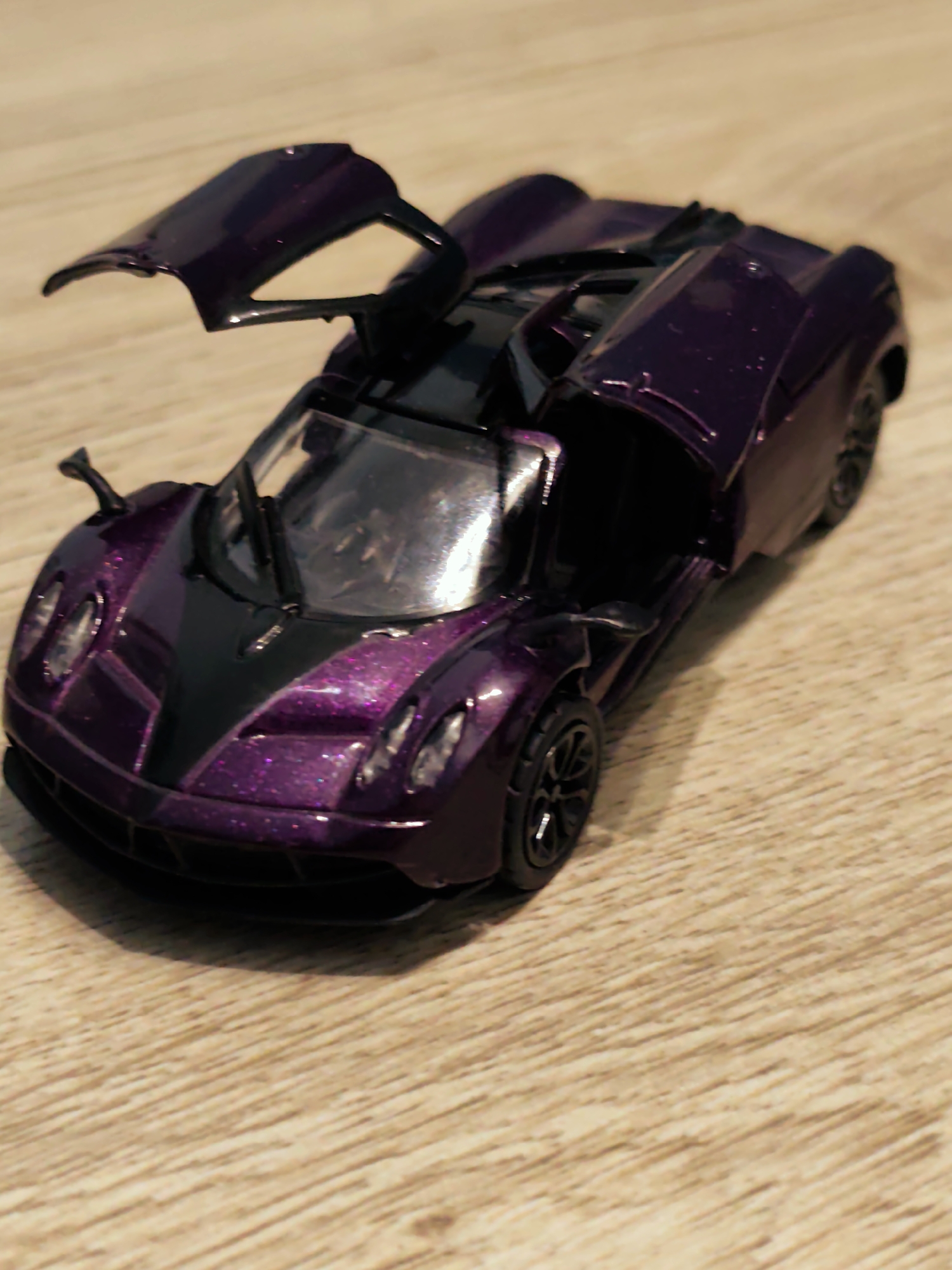 An indoor photograph displays a die-cast model car resting on a light brown wooden surface. The car, a replica of a high-performance vehicle, features a metallic dark purple finish with black accents on the hood and interior components. Both butterfly-style doors are open and elevated. Visible details include black multi-spoke wheels, a left side mirror, and windshield wipers. The wooden surface exhibits distinct linear grain patterns. The scene is illuminated by an unseen light source, casting subtle shadows to the left and rear of the vehicle. No human subjects or additional objects are present. This scene is situated in the city of Pazardzhik, Bulgaria.