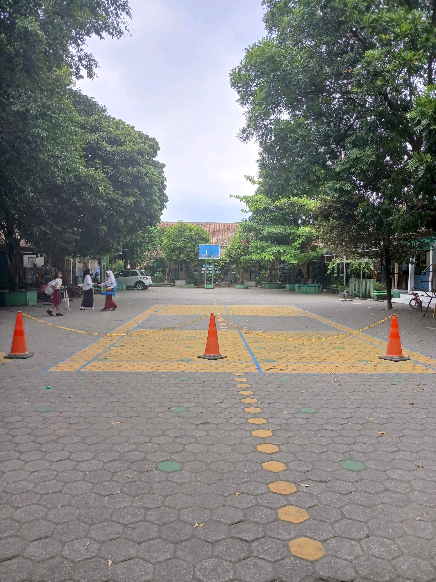 The image depicts an outdoor courtyard at a school in Depok, Indonesia, likely a sports court or assembly area. The ground is paved with hexagonal interlocking concrete blocks, interspersed with some green hexagonal shapes. A yellow painted path with a line of yellow hexagonal markers leads diagonally across the court. Several orange traffic cones are strategically placed, with a yellow chain strung between two of them, suggesting a temporary boundary or queue.
In the background, a white SUV is parked. Three young individuals, likely students, are present. One in a white shirt and dark skirt stands to the left, while two others in white shirts and headscarves, one with a dark skirt and the other with a red skirt, interact in the center. Behind them are buildings with terracotta roofs and trees providing shade. A blue basketball hoop is visible in the distance. A red bicycle is leaning against a railing to the far right. The sky is overcast.