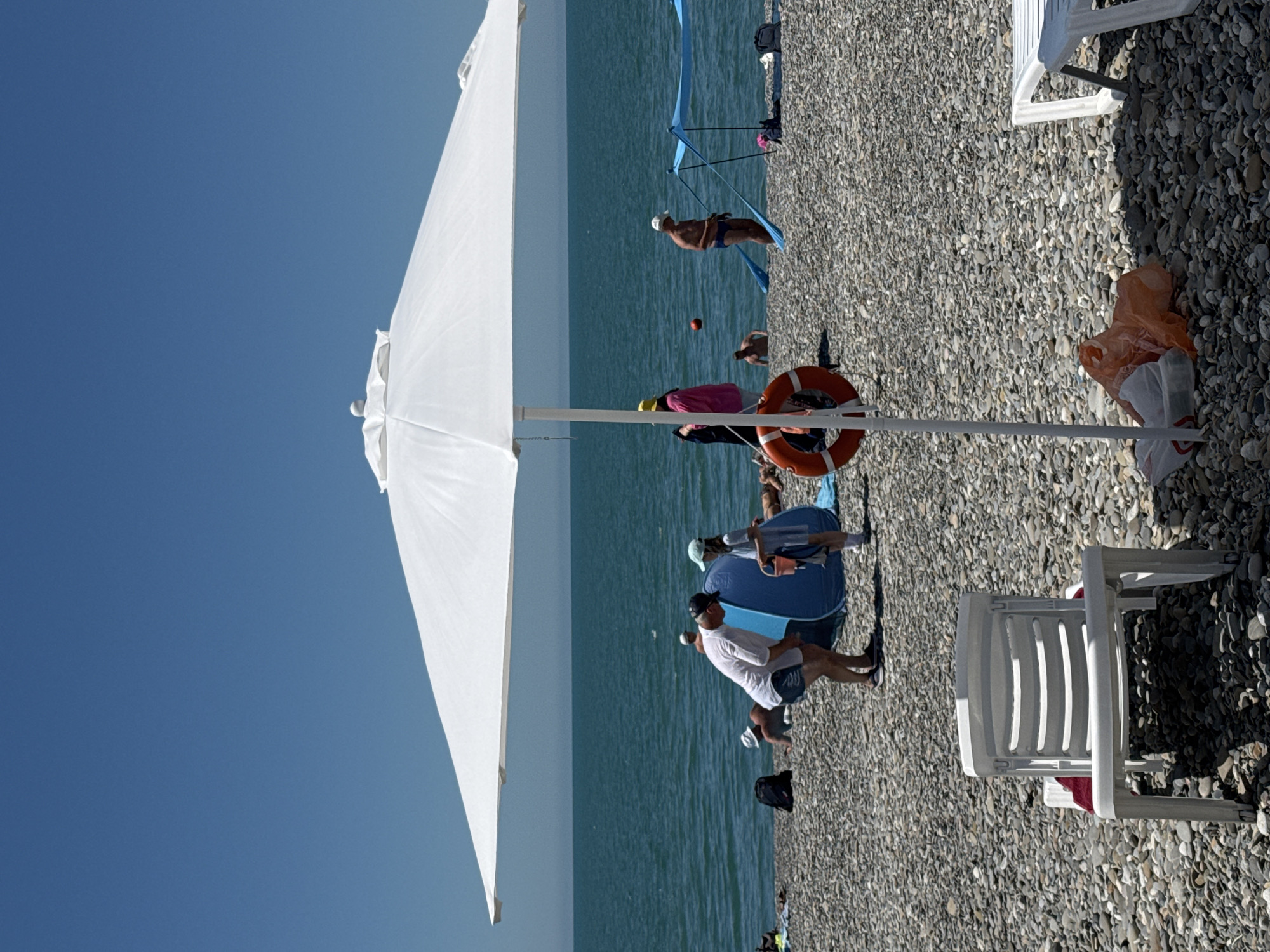 This image captures a wide shot of a pebbled beach in Sochi, Russia, under a clear blue sky on a sunny day. In the immediate foreground, a large white beach umbrella dominates the lower half of the frame, with its central pole supporting a red lifebuoy. To the left of the umbrella pole, a white plastic beach chair is positioned, facing the viewer. To the right, part of another white plastic lounge chair is visible. Below the umbrella and beside its pole, an orange plastic bag and a white plastic bag rest on the pebbles.
In the mid-ground, a man wearing a white t-shirt, dark shorts, and a dark baseball cap is bending forward, looking into a blue, conical pop-up beach tent. A child, dressed in a light-colored top and cap, stands to the man's right, holding a red bucket. Further left, a person is partially visible near a black backpack. In the shallow, turquoise-blue sea, several individuals are present: a person wading waist-deep to the left, another person in blue swim trunks and a white cap standing near a blue fabric sunshade structure to the right, and a red ball floating near another person further out. Another individual in a pink top and yellow hat stands by the water's edge to the left. On the beach, a person is lying down under the blue fabric sunshade, and other figures are visible in the background. The horizon line is visible where the calm sea meets the clear blue sky. The entire beach surface consists of small, grey and brown pebbles.