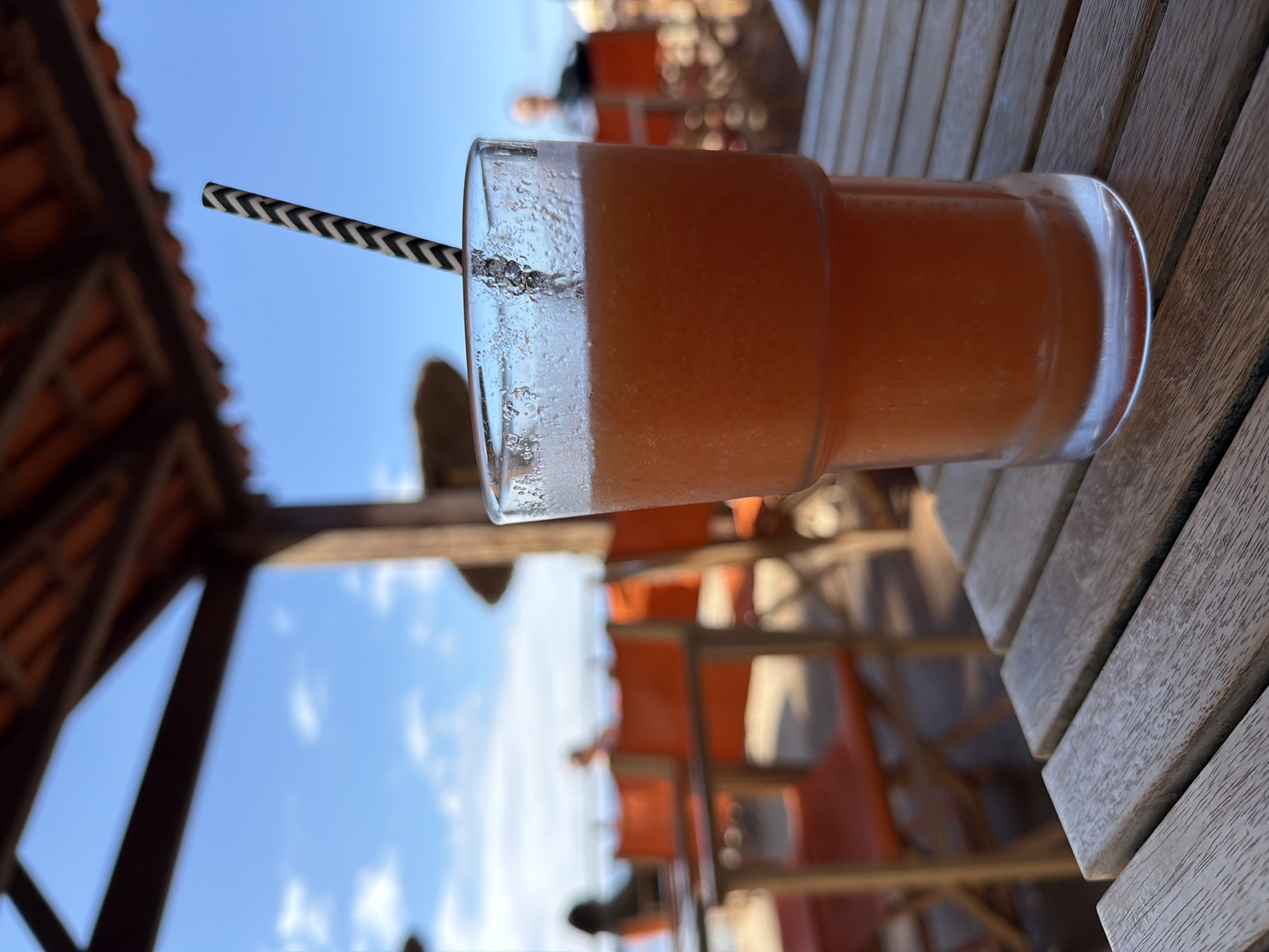 A glass of orange juice with a straw sits on a wooden table. The sky is visible in the background, and the top of a building structure is visible in the left side of the frame. A restaurant patio with orange chairs can also be seen. This scene is likely in Funchal, Portugal.