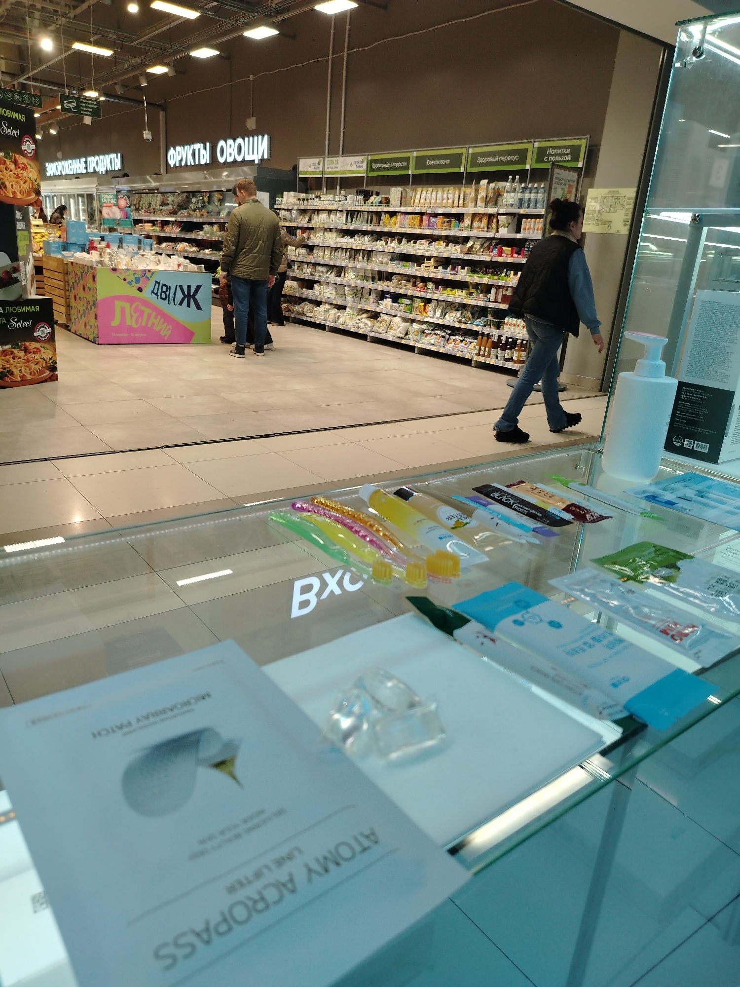 The image captures a scene inside a supermarket in Izhevsk, Russia. The focus is on a display case filled with beauty and personal care products, including items like toothbrushes, creams, and a product labeled "ATOMY ACROPASS LINE LIFTER" with "MICROARRAY PATCH". The case is made of glass.
Beyond the display, the supermarket setting is evident. There are sections labeled in Russian: "ЗАМОРОЖЕННЫЕ ПРОДУКТЫ" (Frozen Products), "ФРУКТЫ ОВОЩИ" (Fruits and Vegetables), "Правильные слодости" (Correct sweets), "Без глютена" (Gluten-free), "Здоровый перекус" (Healthy snack), and "Напитки с пользой" (Drinks with benefits).
Two people are visible: one man in a green jacket and jeans is examining products on a shelf, and another person is walking away in a denim shirt and black vest.
