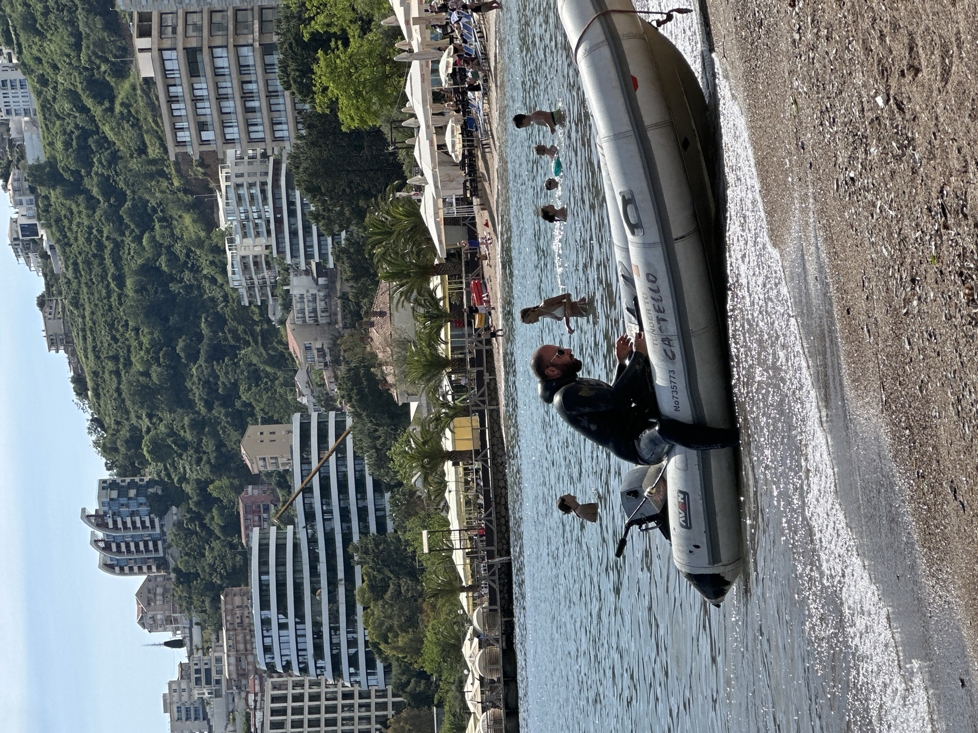 A sideways shot shows a man wearing a black wetsuit sitting in a grey inflatable boat partially on the sand and partially in the water. The boat has the words "TENDER TO CASTELLO" and "No.735773" written on it. The water is clear and reflects the sunlight. In the background, people are swimming and sunbathing on a beach with white umbrellas and palm trees. Further in the background, there are buildings on a hillside covered with trees. The location is likely within Budva, Montenegro, given the architecture and geography.