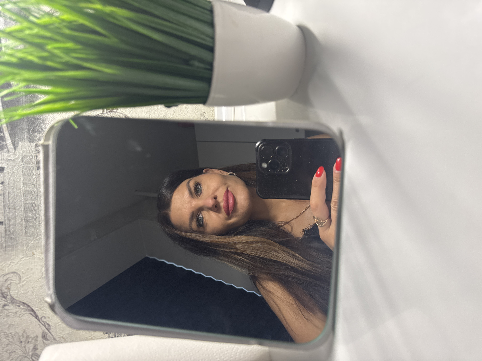 A woman with long, brown hair and blue eyes, wearing red lipstick, is reflected in a mirror. She is holding a black smartphone and wearing a gold ring on her left hand. She also has bright red nail polish on her fingernails. The background is a wall with a textured, patterned wallpaper. To the right of the mirror, there is a white pot with a green artificial plant. The setting appears to be indoors, possibly a bathroom or vanity area. There is no information in the image to suggest it is located in Chaikovsky, Russia.