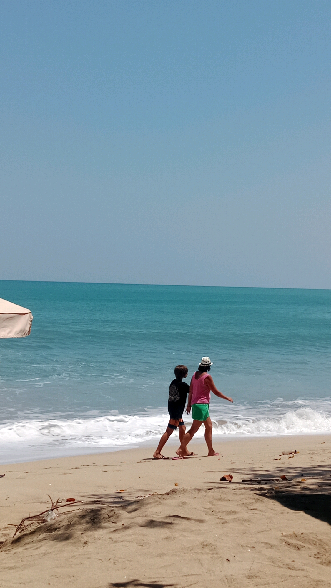 Two individuals, a child and an adult, walk along the sandy shore of Kuta Beach in Indonesia. The child, wearing a black t-shirt and dark shorts, walks slightly ahead of the adult. The adult, dressed in a pink tank top and green shorts, wears a white hat. They are walking in profile, facing away from the viewer, towards the right of the frame. The ocean is a vibrant turquoise with gentle waves rolling onto the beach. The sky above is a clear, pale blue. A portion of a beige beach umbrella is visible on the left side of the image. The sand in the foreground is textured with small debris and shadows cast by unseen objects.