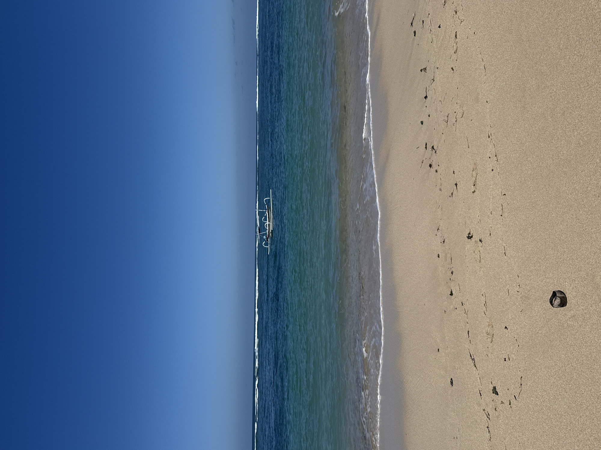 An aerial view of a beach in Kuta Selatan, Indonesia, shows a serene tropical scene. The clear blue sky stretches across the top of the frame, meeting the calm, turquoise waters of the ocean. A traditional Balinese outrigger boat with a white hull and outriggers floats gently on the water, its silhouette distinct against the vibrant blue. The shoreline is a band of light beige sand, with gentle waves lapping at the edge, leaving a thin line of white foam. Scattered across the sand are small pieces of dried seaweed and some footprints, suggesting recent human activity. In the foreground, near the bottom of the frame, a small, dark, rounded object, possibly a shell or a rock, rests on the sand. The overall impression is one of tranquility and natural beauty.
