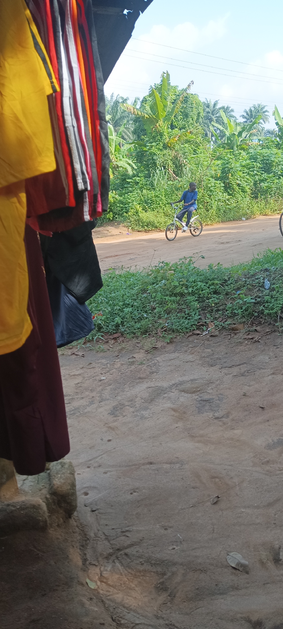 The photograph captures a scene in Amauzu, Nigeria. In the foreground, clothing items in yellow, red, white, and black hang partially obscuring the view. Beyond, a dirt road runs horizontally across the frame, lined with green vegetation. A person wearing a blue shirt rides a bicycle along the road. The background features dense green foliage, suggesting a lush environment. The sky is partly cloudy.