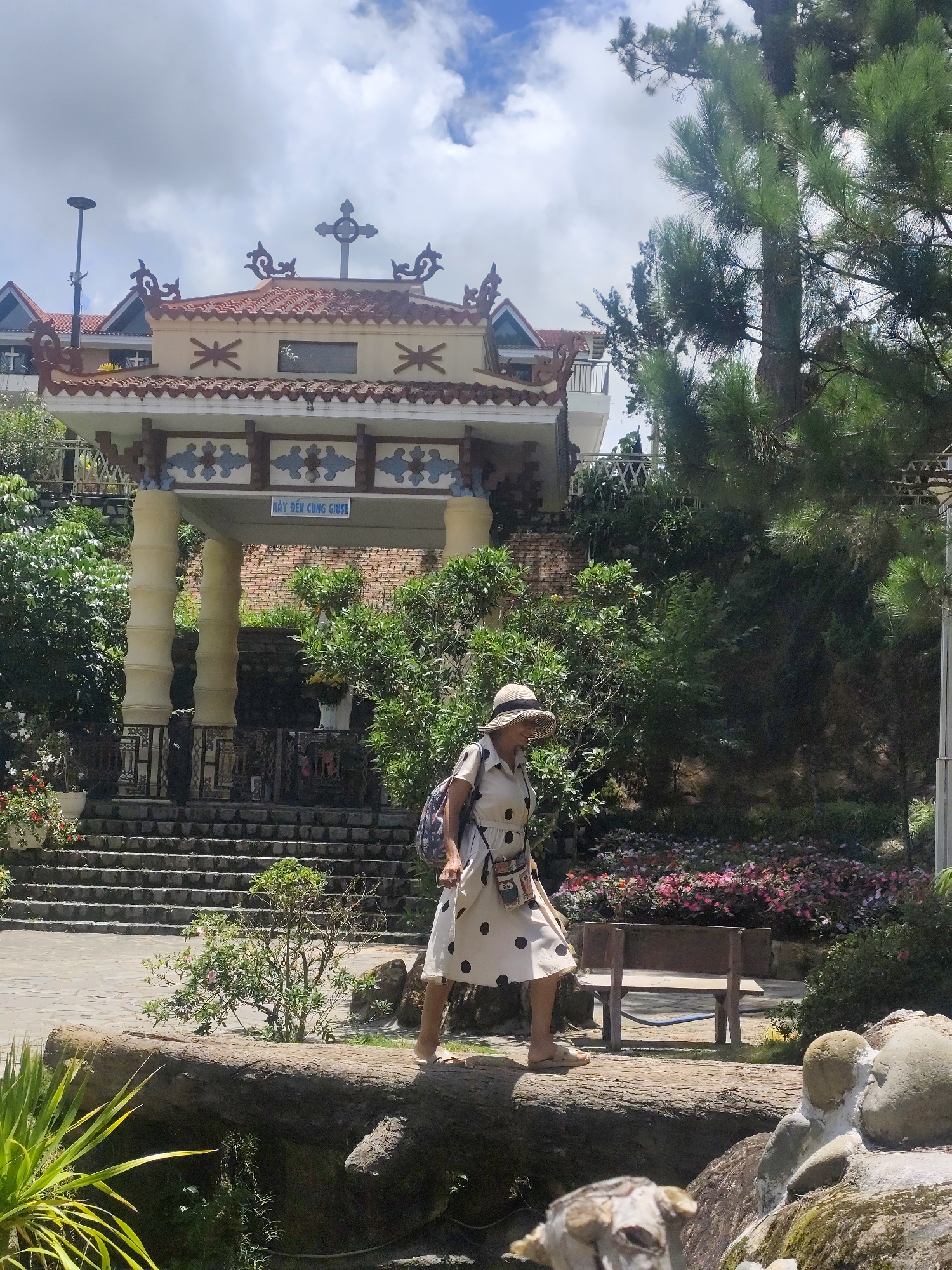 A woman wearing a polka dot dress and a straw hat walks on a fallen log in a garden in Da Lat, Vietnam. The garden features lush greenery, flower beds, and a stone pathway. In the background, a building with a red tiled roof and a cross on top is visible, likely a church or a religious site, given the sign that reads "HÃY ĐẾN CÙNG GIUSE". The sky is cloudy.