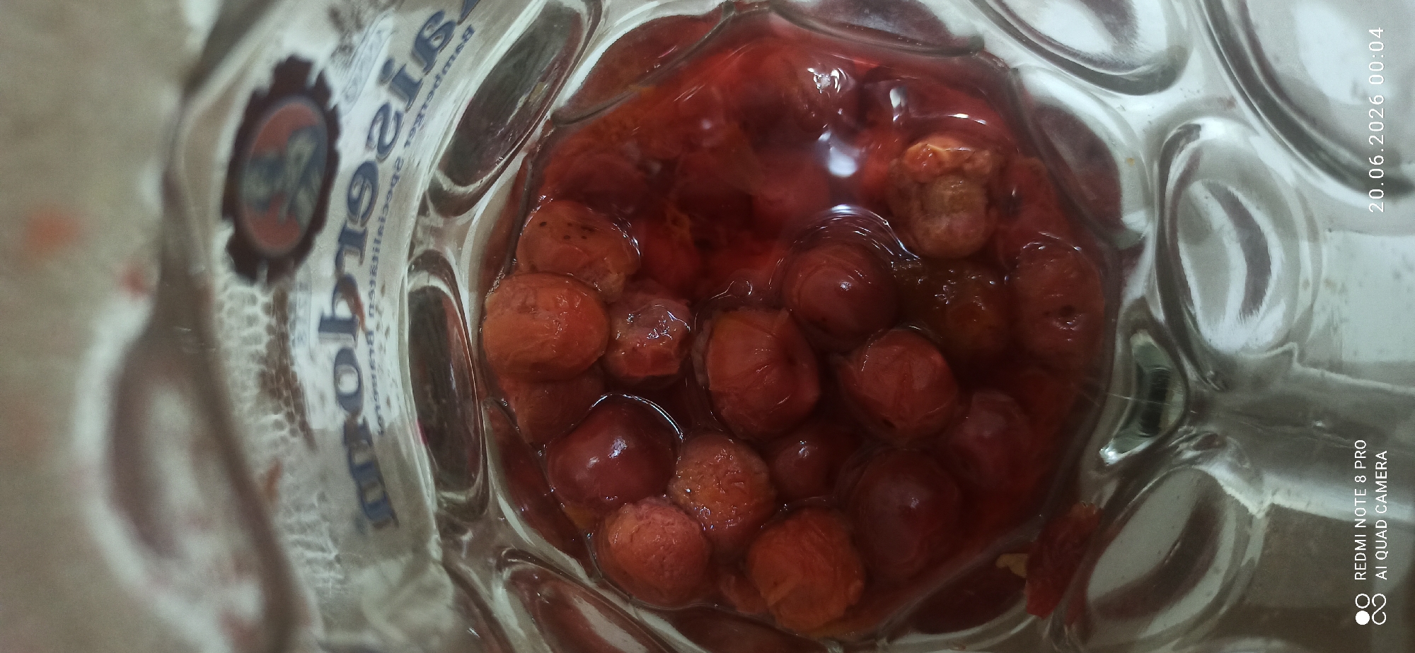 This media file is a close-up, overhead shot of a glass jar filled with whole cherries suspended in a red liquid, likely syrup or juice. The cherries are a deep red to reddish-brown color, with some appearing slightly shriveled, indicating they may be preserved or cooked. The liquid is translucent and appears to be of a syrupy consistency.
The glass jar has a distinctive textured surface with vertical ridges that create a rippled effect on the glass. The lighting is somewhat diffused, but highlights on the glass and cherries suggest an indoor setting. Some of the glass has a label with blue and white lettering, but it is partially obscured and out of focus, making the text unreadable. There is also a circular emblem with a dark outline and a lighter image inside, possibly a crest or logo, also blurred.
Visible text in the bottom right corner of the image provides metadata: "20.06.2026 00:04" and "REDMI NOTE 8 PRO AI QUAD CAMERA". This indicates the image was captured on June 20, 2026, at 00:04 AM, using a Redmi Note 8 Pro phone. The scene is likely a kitchen or pantry, given the presence of preserved fruit in a jar. No people or explicit weather conditions are discernible. The overall impression is one of preserved fruit, possibly homemade jam or compote.