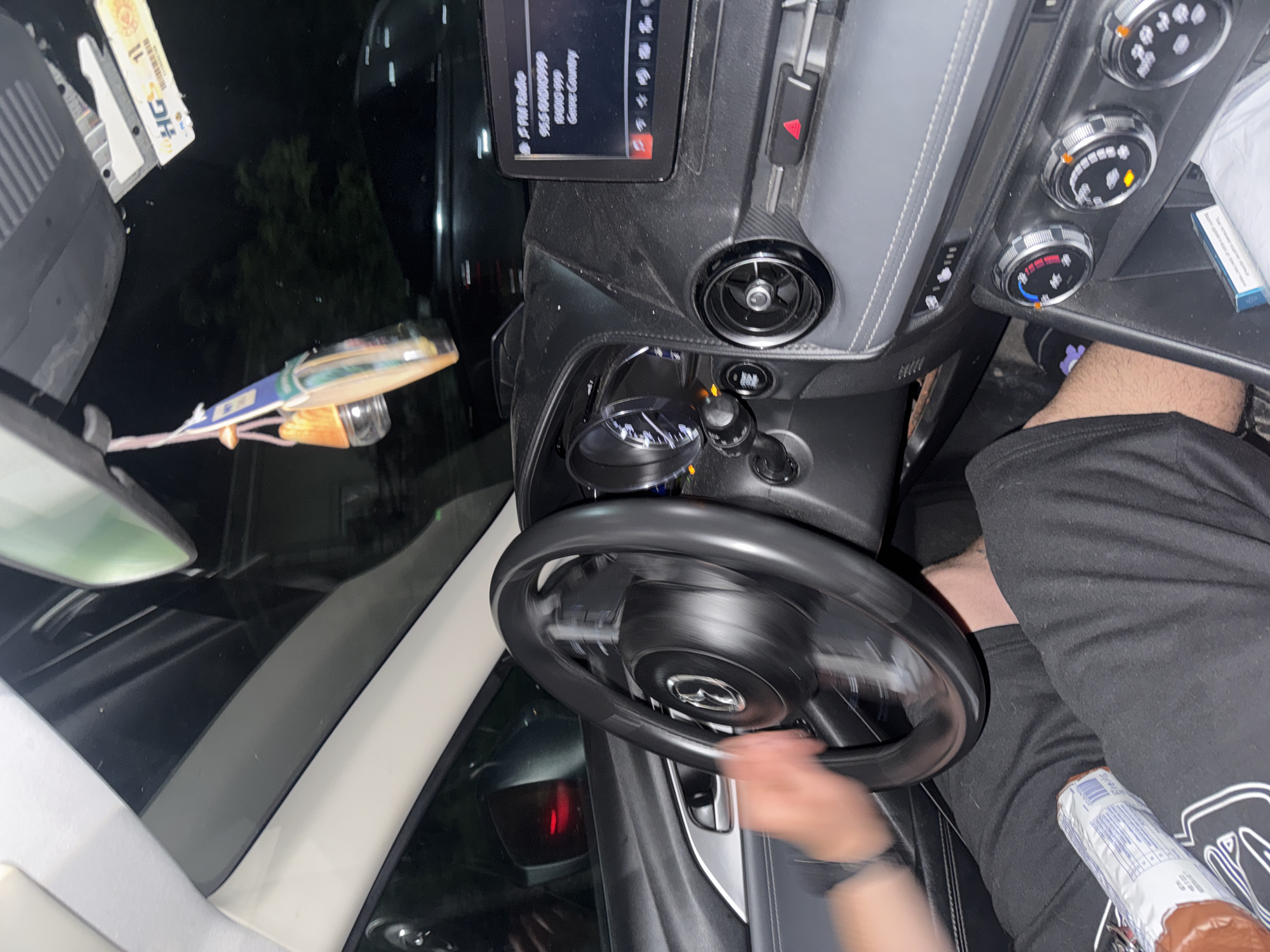The image shows the interior of a car. A person is holding the steering wheel. The car's dashboard includes a screen displaying a radio station frequency and name, as well as climate control dials. A packet is also visible on the passenger's side. The location of the scene is unknown.
