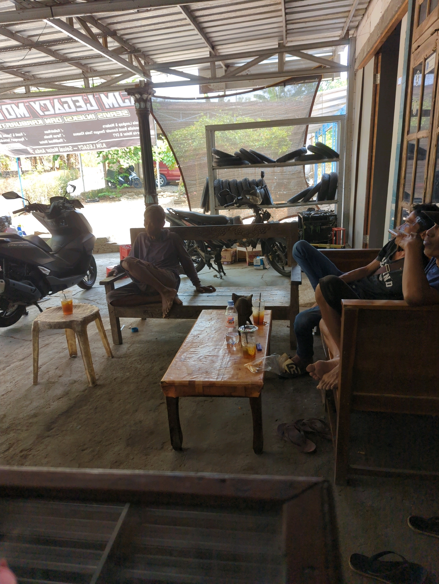 The image depicts an outdoor setting, likely a workshop or garage, in Kedungdowo, Indonesia. In the foreground, a man is seated on a wooden bench, smoking a cigarette. Another man is sitting on a chair to his left, appearing to be engaged in conversation or observing. In the background, several motorcycles are parked, suggesting an automotive repair shop or dealership. Shelves behind the motorcycles are filled with spare tires. The overall environment appears to be somewhat cluttered but functional.