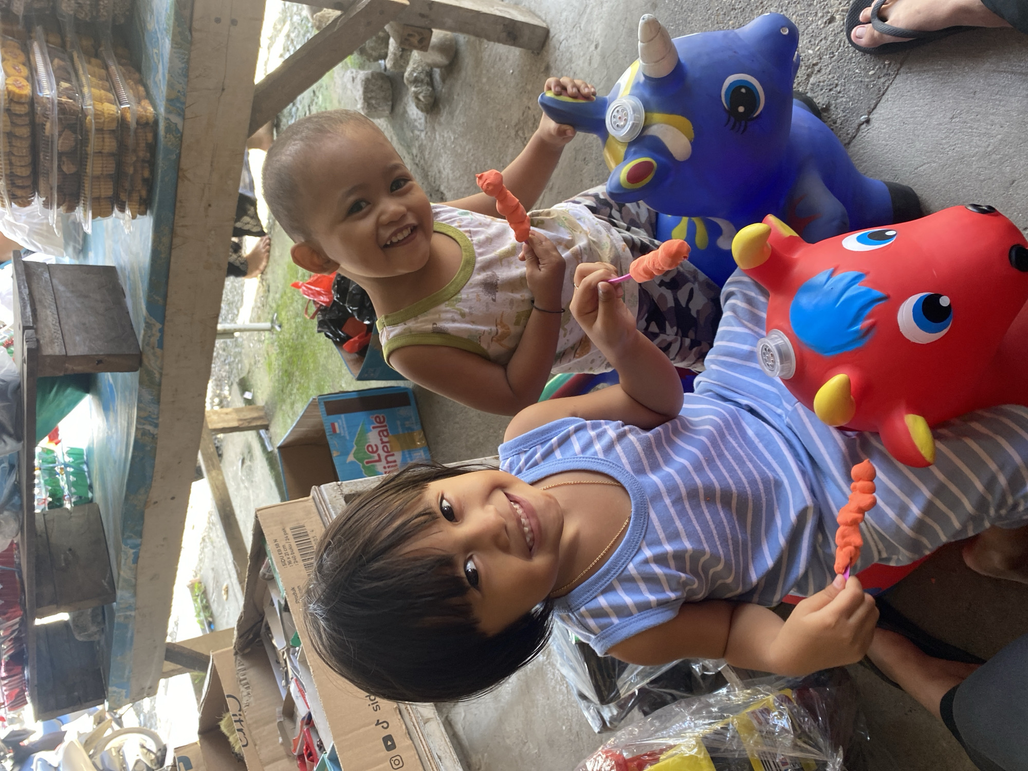 Two young children, a girl and a boy, are playing with orange modeling clay while sitting on inflatable animal toys. The girl, on the left, wears a blue and white striped sleeveless shirt and a gold necklace. Her dark hair is cut short and styled with bangs. The boy, on the right, wears a white tank top with a green trim and camouflage shorts. Both children are smiling. The girl is holding a piece of orange clay on a pink stick. The boy is holding a similar piece of clay. They are sitting on a red and a blue inflatable toy animal, possibly horses or unicorns.
The environment appears to be an outdoor market or roadside stall. In the background, there are wooden tables with various items, including stacks of cookies in clear plastic containers and boxes containing goods. One box has a label with "Le minerale" text. The ground is concrete. The overall impression is of a casual, possibly rural or semi-urban setting in Ta', Indonesia. The lighting suggests it is daytime, possibly under some cover from the sun.