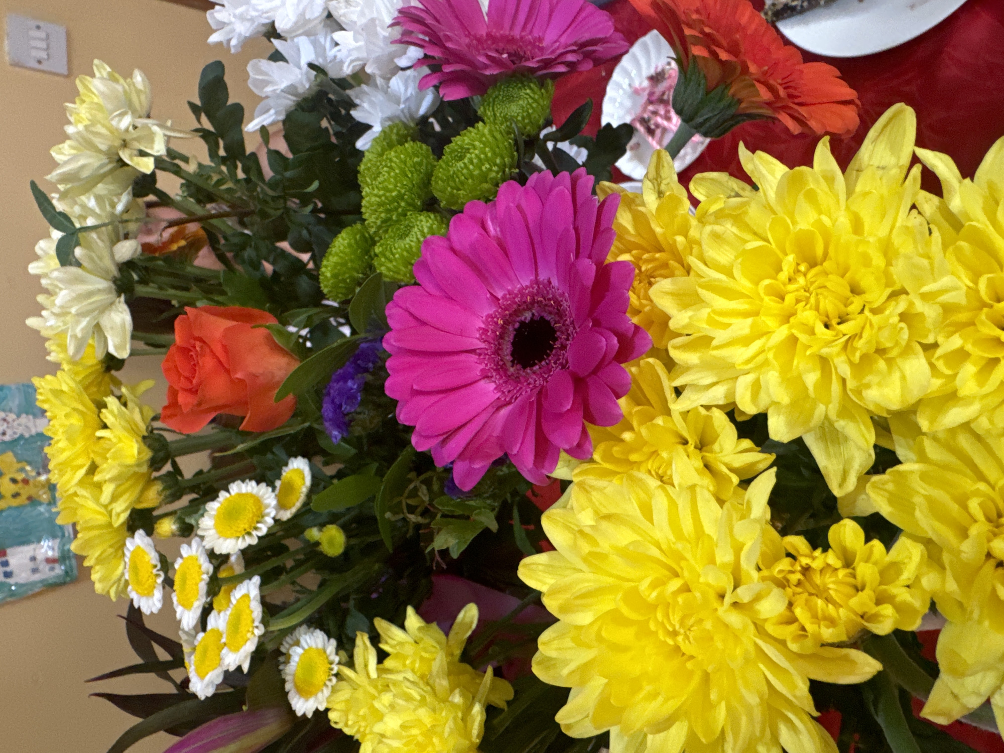 The image shows a close-up, overhead view of a vibrant bouquet of flowers arranged in a bowl on a red surface. The bouquet is a mix of chrysanthemums and gerbera daisies in various colors, including bright yellow, hot pink, white, and orange. There are also small, white daisy-like flowers with yellow centers, and some small green pom-pom like flowers. In the background, a framed drawing hangs on a wall, and a white electrical outlet is visible. The lighting suggests an indoor setting. There is no indication of the city of Waterford, Ireland, or any specific location within it.