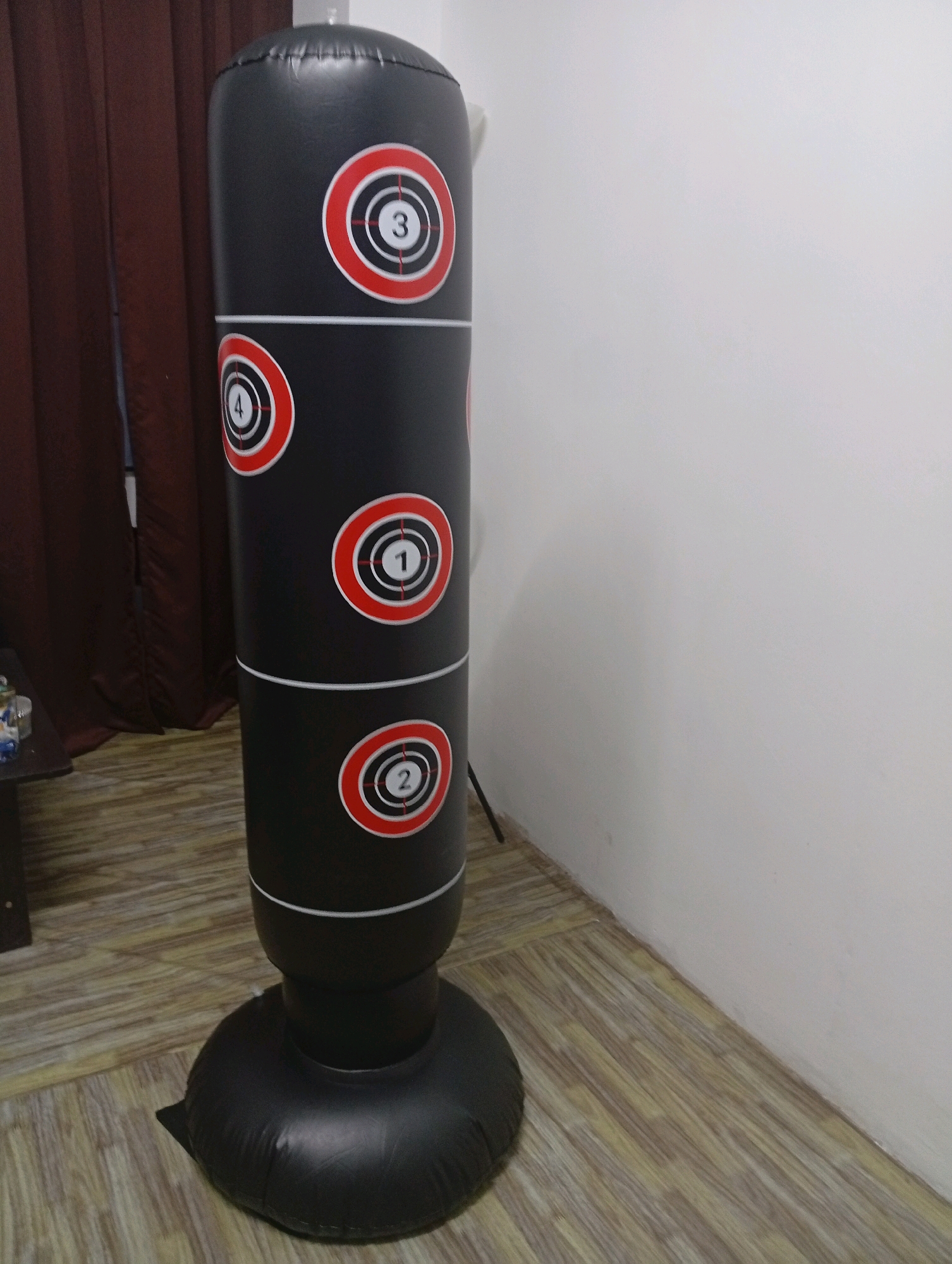 An inflatable black punching bag with red and white circular targets is standing on a wooden floor indoors. The punching bag has targets labeled "3", "4", "1", and "2" from top to bottom. The bag is cylindrical and has a weighted base. To the left of the punching bag, there is a dark brown curtain. The wall behind the punching bag is white. The image appears to have been taken in a residential or recreational setting. The specific location in Selayang Municipal Council, Malaysia, cannot be determined from the visual information provided in the image.