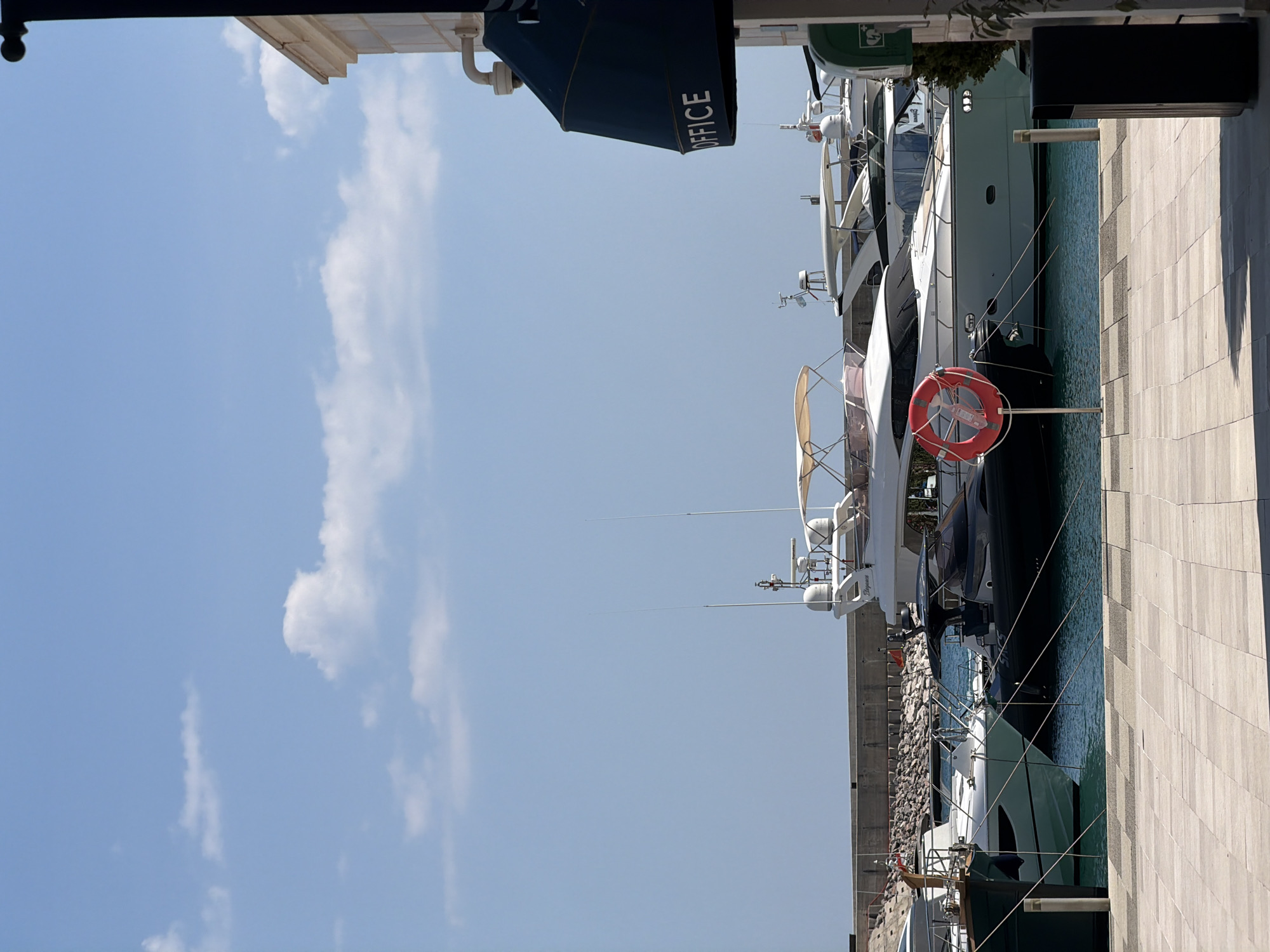 This image shows a view of a marina in Radovići, Montenegro. Several yachts are docked along a paved walkway. The sky is clear blue with scattered white clouds, suggesting fair weather. The time of day appears to be daytime, likely morning or afternoon, given the lighting.
The yachts vary in size and design, with some appearing modern and luxurious. One yacht has a dark hull, while others are white. Visible on the side of one of the yachts, partially obscured by a dark blue structure, is the word "OFFICE" in white lettering. A red life preserver hangs from a stand on the dock, a common safety feature in such locations. The walkway is made of light-colored, square paving stones. Beyond the yachts, a rocky embankment is visible, and a building with awnings can be seen in the upper left corner. The water in the marina is a calm, greenish-blue. The overall scene conveys a sense of leisure and maritime activity.