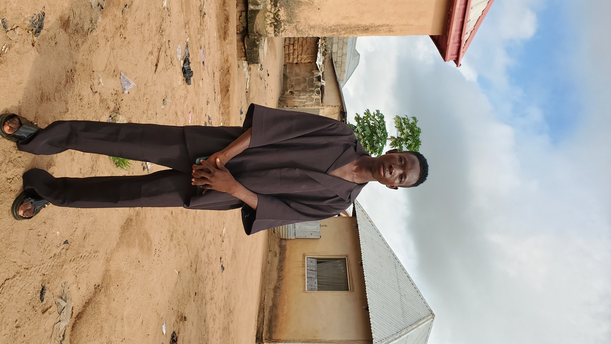 A young Black male stands facing forward in the center of the frame. He is wearing a loose-fitting, dark brown collared shirt with wide sleeves, and matching dark brown pants. He has on black slide sandals. His hands are clasped in front of him. He is standing in what appears to be a rural or undeveloped area. The ground is dry, sandy earth with some scattered debris. In the background, there are several small, simple buildings with corrugated metal roofs and stucco or mud walls. A leafy green tree is visible behind and to the left of the man. The sky above is cloudy with patches of blue sky visible. The overall scene suggests a hot, possibly humid climate. The location is described as Unknown, Unknown.