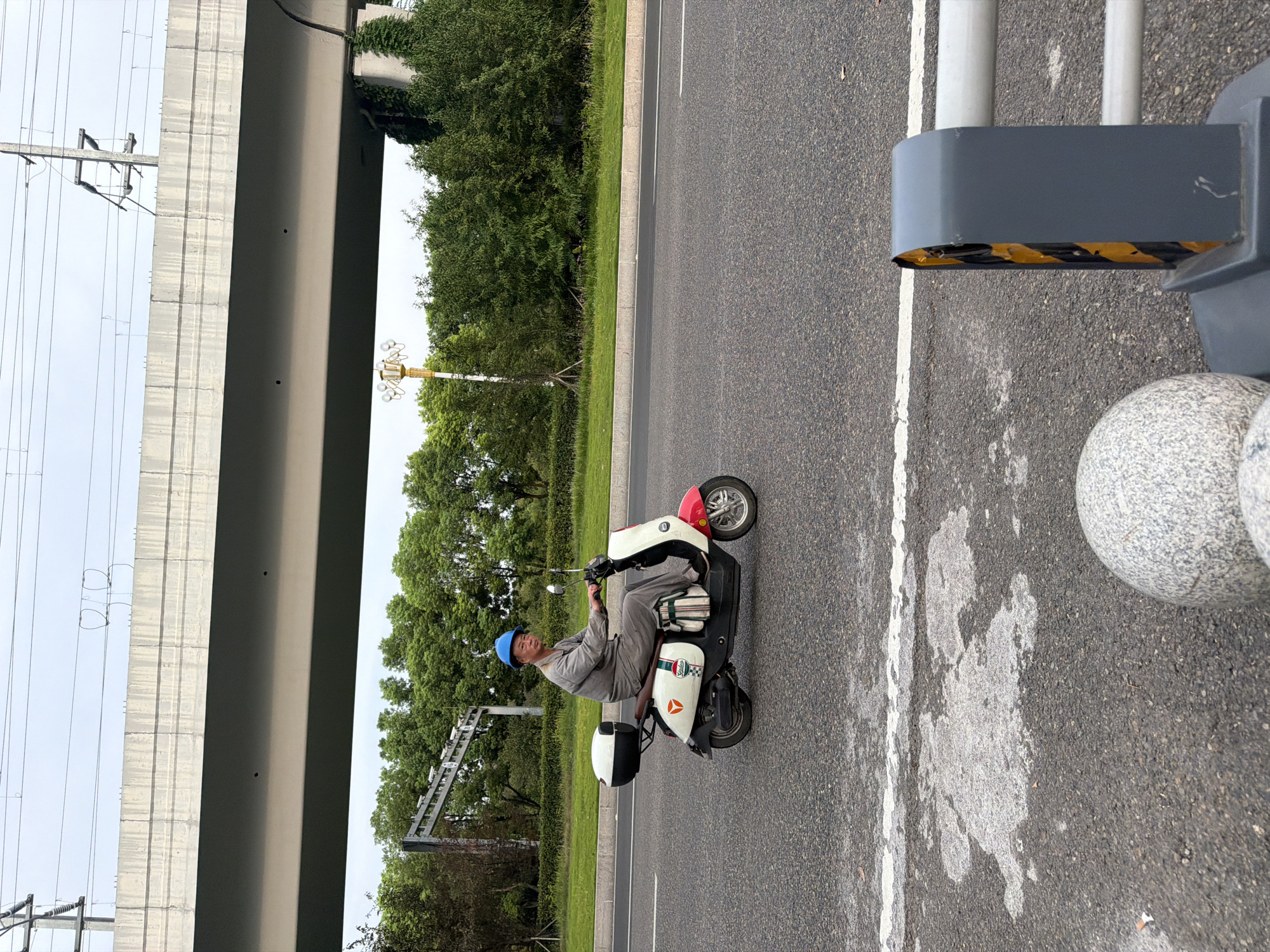 A man wearing a blue hard hat and grey work clothes rides a white and red electric scooter on a paved road. He is looking towards the camera. The scooter has a black storage box on the back and a striped bag hanging from the side. In the background, there are lush green trees, a large concrete overpass, and overhead power lines. The road has white lane markings and some discoloration. In the foreground, a grey concrete bollard and two spherical concrete objects are visible. The scene appears to be located in Jiangjin District, Chongqing, China, given the typical urban infrastructure and vegetation.