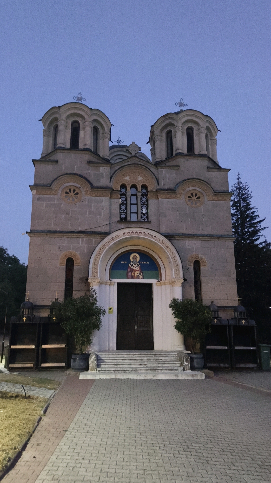 The image shows the exterior of a stone church with two domes topped with crosses. The church is made of light-colored stone blocks and features arched windows and decorative circular motifs. A large, ornate archway above the entrance displays a religious icon of Saint George. Two dark wooden doors are set within this archway. To the sides of the entrance are two potted trees. In front of the church, a wide set of stone stairs leads up to the entrance. A paved pathway, made of interlocking grey and red bricks, forms the foreground. The sky above is a clear, pale blue, indicating dusk or dawn. The church is situated in a slightly elevated position, with some trees visible on the right side of the building. To the left and right of the church entrance are dark, open structures with shelves. A green trash bin is visible to the far right of the image. The overall scene is peaceful and austere. This is the Saint George's Church in Leshok, North Macedonia.