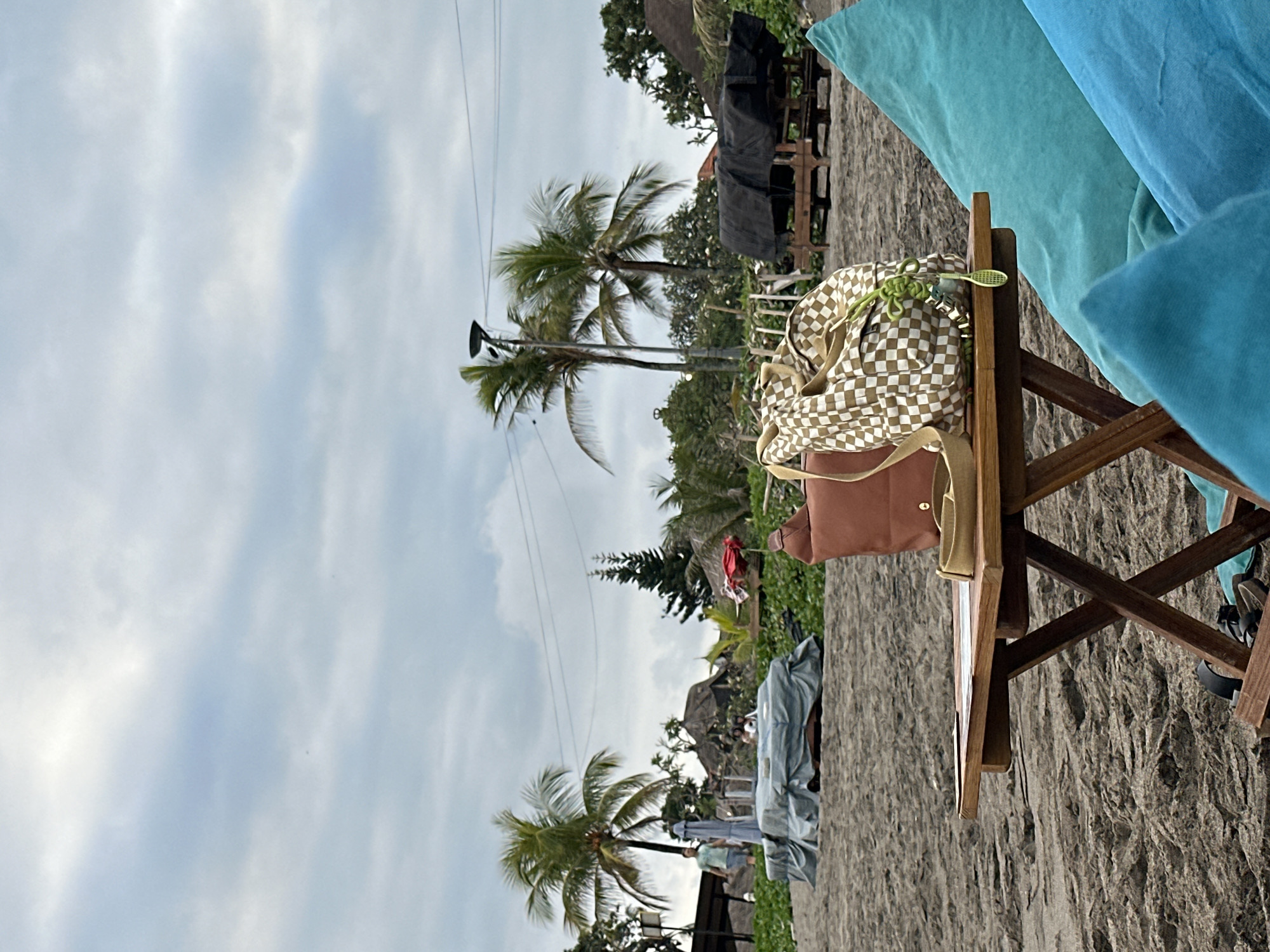 The image depicts a beach scene in North Kuta, Indonesia, under a cloudy sky. In the foreground, a small wooden table with criss-crossed legs sits on the sand. On the table are two bags: a brown leather bag and a checkered patterned bag. A small green decorative item, resembling a tiny tennis racket, hangs from the checkered bag. To the right of the table, large turquoise beanbag chairs are visible. The background reveals a sandy beach with scattered vegetation, palm trees, and thatched-roof structures. Some tables covered with tarps are also present. Two people are visible in the mid-ground walking on the beach. The overall atmosphere suggests a relaxed beachside setting.