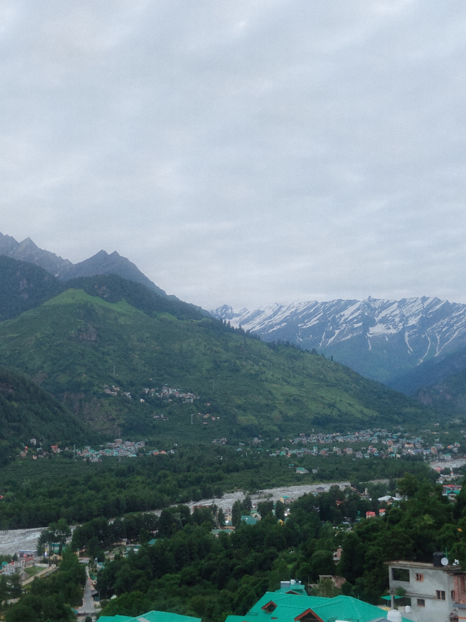 This image captures a panoramic view of Vashisht, India, under an overcast sky. The scene is dominated by lush green mountains that recede into the distance. Snow-capped peaks are visible in the far background, contrasting with the verdant slopes below. A small town is nestled within the valley, with buildings clustered together amidst dense foliage. A river flows through the valley, its waters appearing muted under the cloudy sky. The foreground features more of the town, with houses featuring distinctive green roofs, along with trees and some visible infrastructure. The overall impression is one of a serene mountain village.
