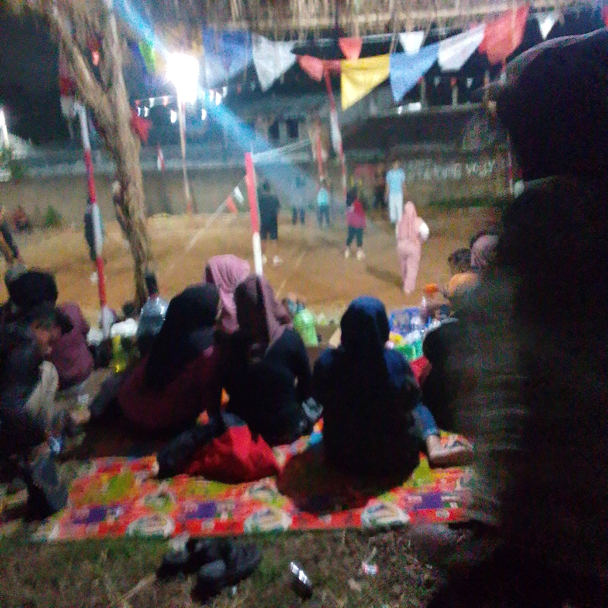In the city of Cilaku, Indonesia, a group of individuals are gathered outdoors at night. The scene is illuminated by artificial lights, casting a soft glow on the surroundings. In the foreground, several people are seated on a colorful mat, engrossed in conversation or observation. One person, in particular, is wearing a dark hooded garment, partially obscuring their face. In the background, more people are visible, some standing and others walking. Colorful triangular flags are strung overhead, adding a festive touch to the atmosphere. The environment appears to be an open area, possibly a park or a community gathering space, with trees and a building visible in the distance. The overall impression is one of a relaxed and social occasion.