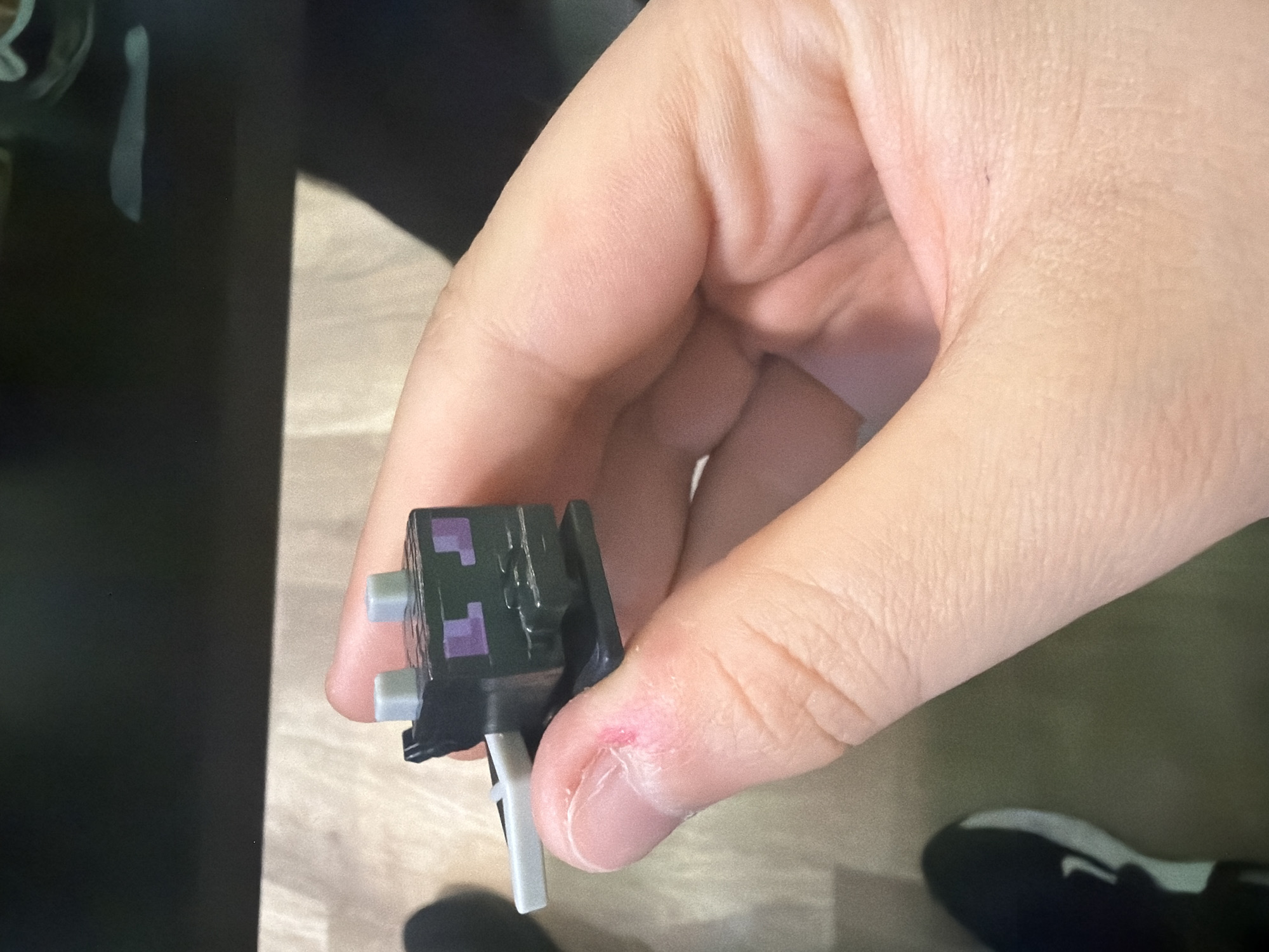 A close-up shot shows a human hand holding a Minecraft Enderman toy. The hand's fingers are curved around the toy, which is black with purple details and gray appendages. The thumb has a small, red mark near the nail. The background is a mix of dark and light surfaces. A black object is visible in the lower right corner of the frame.