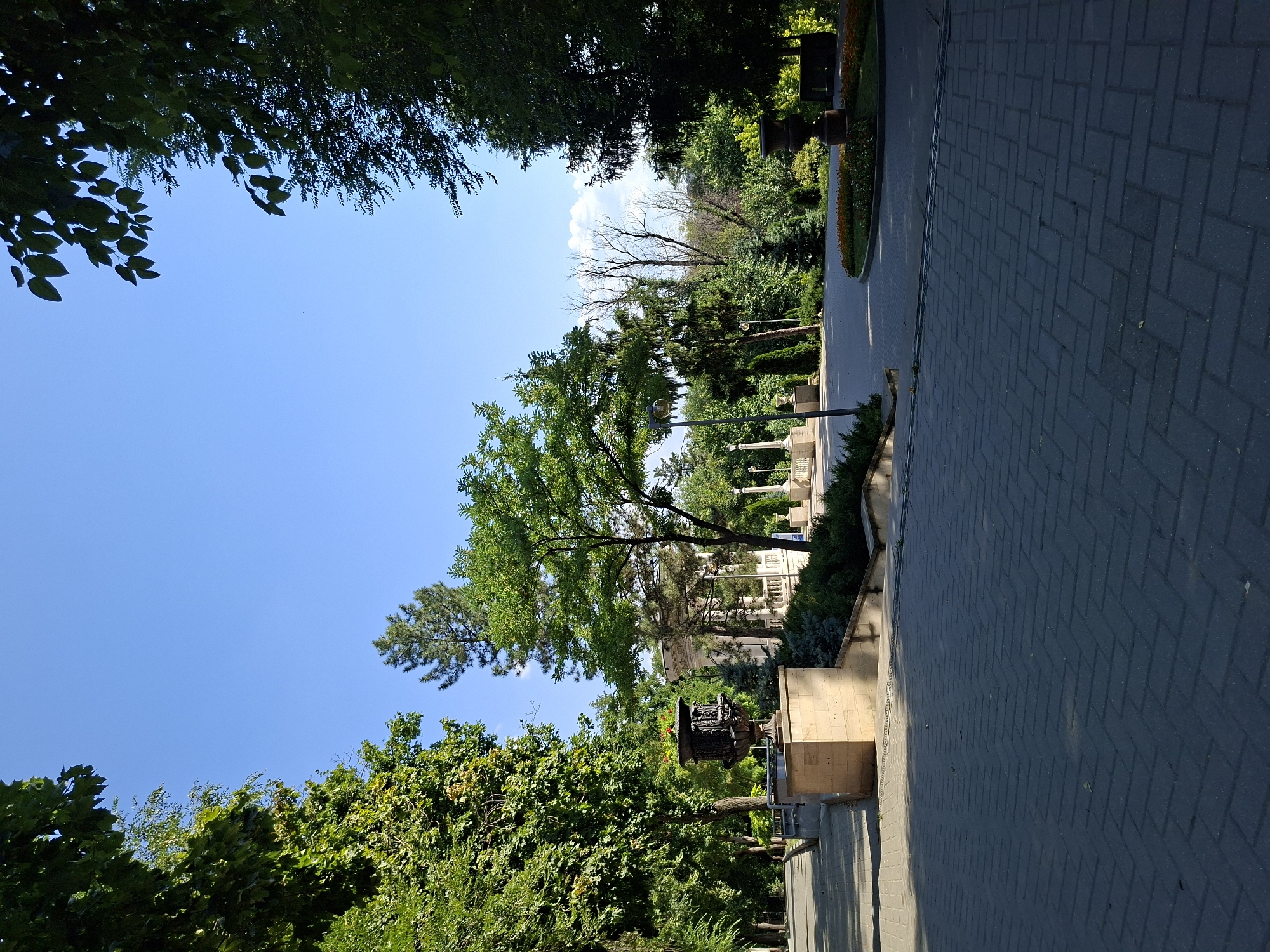 This image captures a serene park scene under a clear blue sky. In the foreground, a paved walkway made of dark gray rectangular stones is laid out in a herringbone pattern. To the left of the walkway, a large, ornate dark metal urn sits atop a light beige stone pedestal. Beyond the urn, a paved path slopes gently upwards and to the left, bordered by lush green trees.
In the mid-ground, a white, classical-style gazebo with columns stands surrounded by manicured bushes and trees. Lampposts with globe lights are visible along the pathways. To the right of the gazebo, a circular flower bed with orange and red flowers is present. Further to the right, a dark urn sits on a low stone base, and beyond that, a dark bench. The background is filled with a variety of trees, some tall and slender, others full and bushy, creating a sense of depth and natural beauty. The overall impression is one of a well-maintained public park, possibly a place for quiet contemplation or leisurely strolls.
This scene is located in Chișinău, Moldova, and depicts a portion of the Stephen the Great Central Park, a prominent green space in the city.