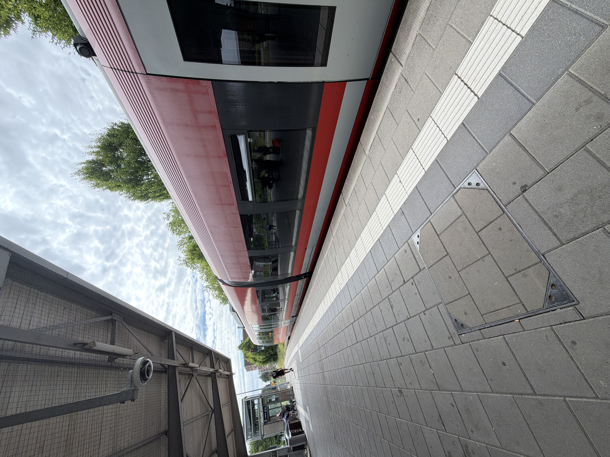 The image shows a train platform at Gronau (Westfalen) station in Gronau, Germany. A red and silver train is stopped at the platform. The platform is paved with grey rectangular tiles and has a tactile paving strip along the edge. A covered walkway with a metal roof extends over part of the platform. A security camera is visible mounted on the underside of the walkway roof. In the background, there are trees and buildings. A few people are visible on the platform, including one person running and another person with a stroller.