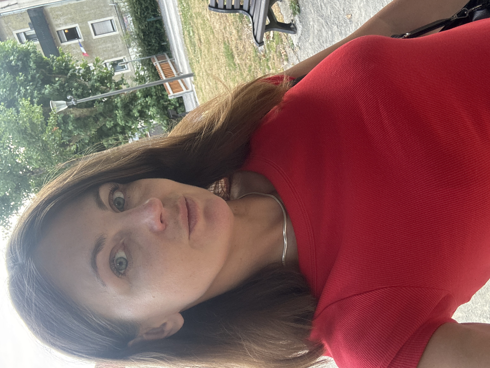 A woman with brown hair and blue eyes is photographed outdoors in a park setting. She is wearing a red top and a silver necklace. Behind her, a building with windows and a balcony is visible, along with trees and a park bench. The lighting suggests it is daytime.