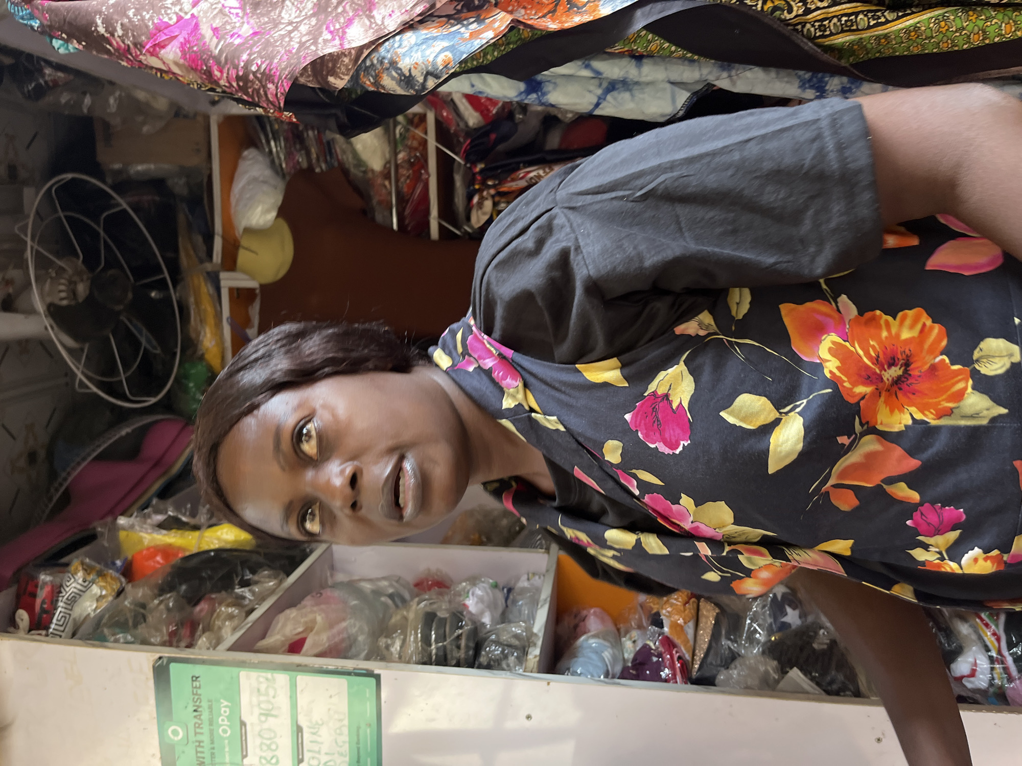 The image shows a woman standing inside a shop, possibly in Abuja, Nigeria. She is wearing a black dress with a floral print of pink, orange, and yellow flowers. The shop appears to be filled with merchandise, with shelves stocked with various items wrapped in plastic. To the left of the woman, a green sign is visible with the text "WITH TRANSFER," "ISTER & MORE RELIABLE," and "OPay." Below this, there are numbers "880 9052" and some handwritten text. A ceiling fan is visible in the background, and fabrics with colorful patterns hang to the right. The overall impression is that of a retail space selling clothing or accessories.