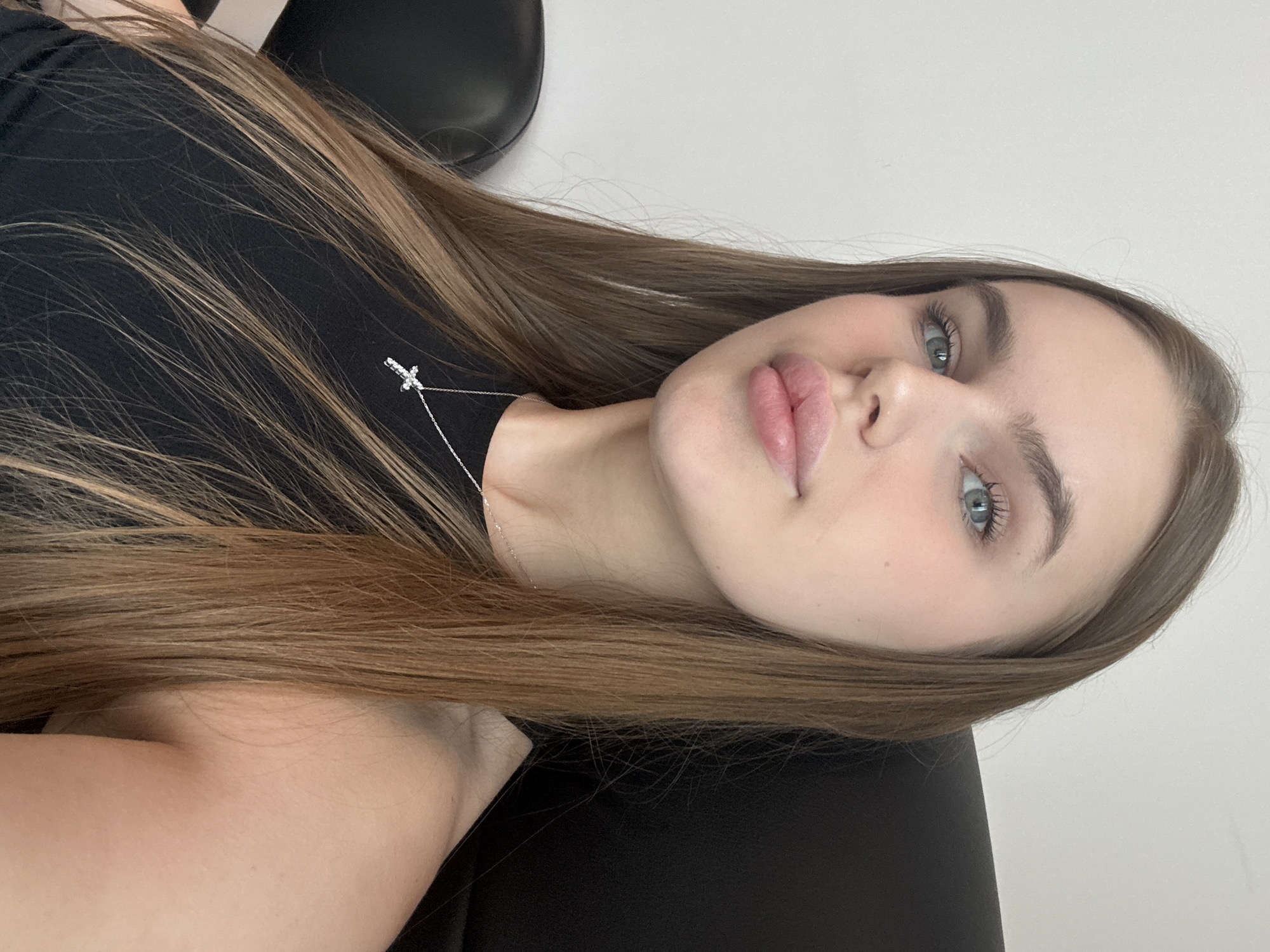 The media file is a close-up, portrait-style photograph of a young woman with long, light brown hair. She is wearing a black top and a delicate silver necklace with a small cross pendant. Her hair is parted on the side and falls across her face and shoulder. The woman's face is turned towards the camera, with her eyes open and looking slightly off to the side. Her makeup appears natural, with a focus on her lips, which are slightly parted and have a glossy finish. She has clear skin and subtly defined eyebrows.
The background is mostly out of focus, but appears to be a light-colored wall or surface on the right side and a dark object, possibly a chair or headrest, on the left. The lighting suggests an indoor setting, with soft, natural light illuminating her face. The overall mood of the image is serene and contemplative. There is no visible text in the media file. The metadata indicates the location context is Dnipro, Ukraine.