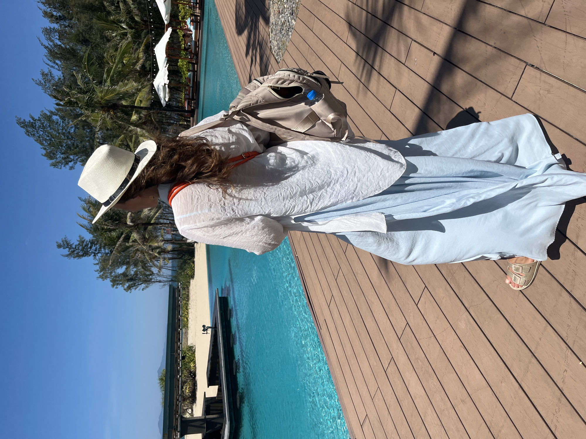 An overhead, slightly angled shot captures a person, seen from behind, standing on a wooden deck beside a swimming pool under a bright sky. The person, appearing to be a woman with long brown hair, wears a white straw hat, a loose-fitting white long-sleeved top, and light blue flowing trousers or a skirt. A beige backpack is visible on her shoulders, with a blue water bottle protruding from a side pocket. An orange strap is visible beneath her white top. Her right foot, wearing a light-colored sandal, is visible on the deck.
The scene depicts a sunny day, with strong shadows of nearby trees cast across the wooden deck. The clear turquoise water of the infinity pool stretches out next to the deck. Beyond the pool, a sandy beach area is visible, lined with numerous palm trees and other green foliage. In the distance, under a clear blue sky, soft outlines of mountains or hills can be seen. The overall setting suggests a tropical or coastal resort environment in Đà Nẵng, Vietnam, conveying a sense of leisure and relaxation. There is no visible text in the image.
