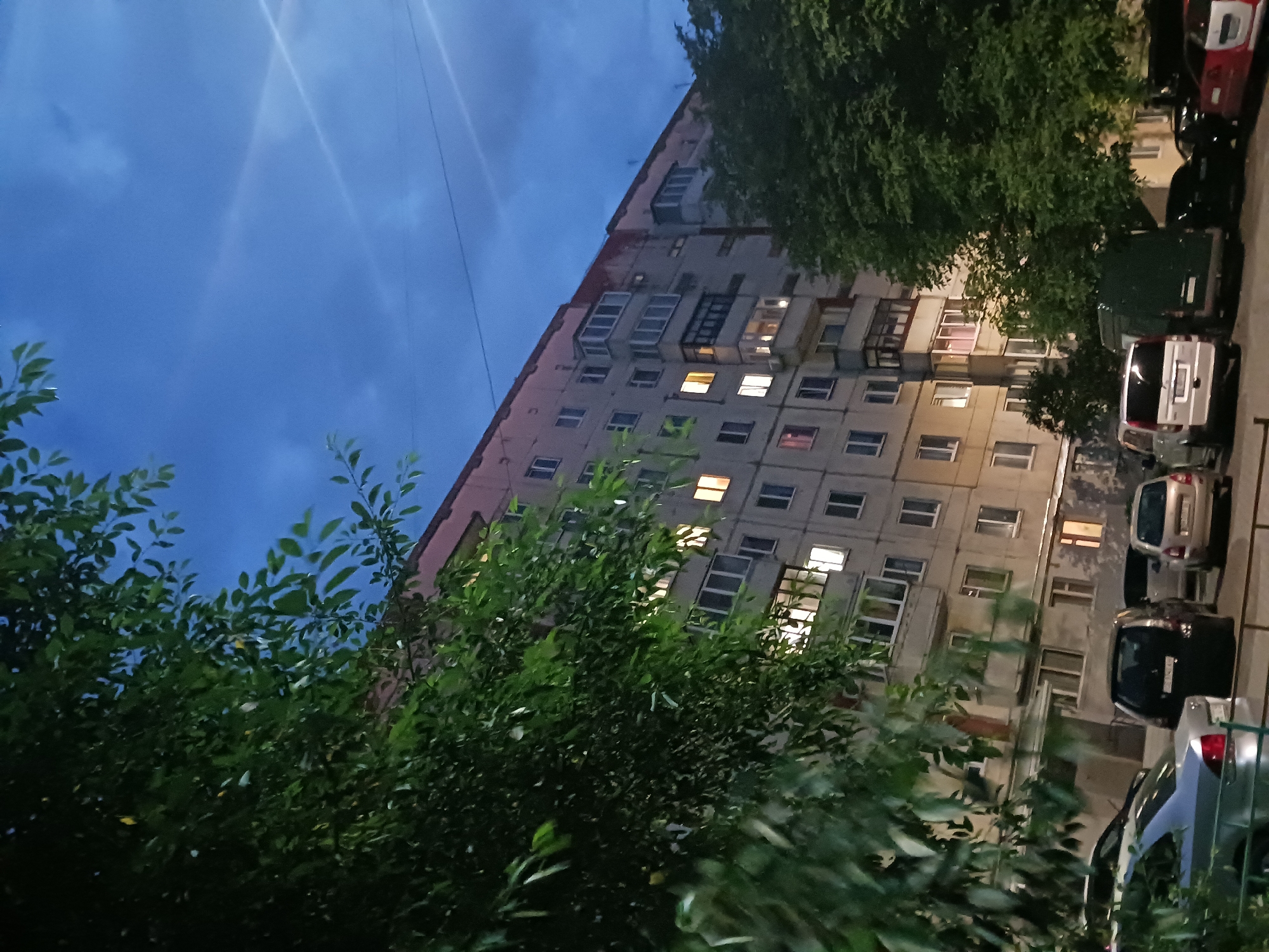 The image depicts a nighttime scene in a residential area, likely in an urban or suburban setting. The primary subject is a multi-story apartment building, its facade illuminated by interior lights visible through numerous windows. Some windows show a warm, yellowish glow, suggesting interior lighting, while others are darker. A pinkish hue is visible along the top edge of the building's facade.
In the foreground and to the left, a large tree with dense green foliage partially obscures the view of the building. The leaves are dark due to the low light conditions. To the right, another tree with similar foliage is visible, also partially obscuring the building.
In front of the apartment building, several cars are parked. These include a silver hatchback, a black minivan or SUV, a white van, and what appears to be a red car. The vehicles are parked in a lot or street in front of the building.
The sky above is a deep blue, indicating it is either dusk or night. There are some faint clouds and what appear to be light streaks or reflections, possibly from artificial lights or the camera's lens. The overall atmosphere is one of a quiet evening in a populated area. The specific location is listed as Unknown, Unknown.