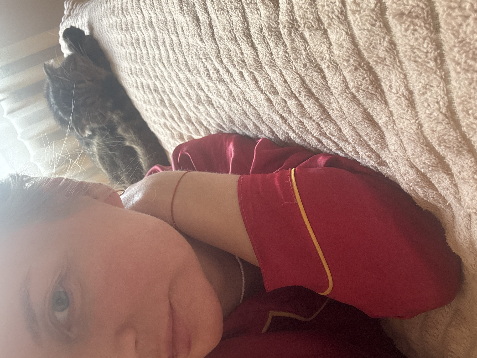 The image shows a person lying on a bed, wearing red pajamas with yellow trim. A grey tabby cat is standing near the head of the bed. The location is not identifiable as the city of Chaikovsky, Russia from the image alone.