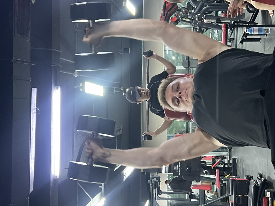 The image captures an indoor gym scene, located in Nha Trang, Vietnam. In the foreground, a Caucasian male with short light-colored hair, wearing a black tank top, lies supine on a red padded weight bench. He is actively engaged in a dumbbell press, holding two large black hex-style dumbbells with his arms fully extended vertically overhead. A tattoo is visible on his right forearm. His gaze is directed towards the camera.
Immediately behind him, an Asian male wearing a dark t-shirt and a dark baseball cap sits on another red padded surface. He holds a small, black, cylindrical weight in each hand, with his arms extended laterally, consistent with a spotting position or a concurrent lateral raise exercise.
The background shows additional gym equipment, including weight machines, one of which is partially labeled "4N1 PEC FLY LATERAL RAISE STRENGTH". A third male, wearing dark clothing and light shorts, is partially visible standing near equipment on the far left. On the far right, the hands and forearms of a fourth individual are partially visible near another piece of equipment. A blue and clear water bottle with a white cap rests on the floor to the right.
The gym features a dark ceiling with exposed ventilation ducts, electrical conduits, and various artificial light sources, including multiple long fluorescent tube lights and rectangular LED panel lights, illuminating the space.