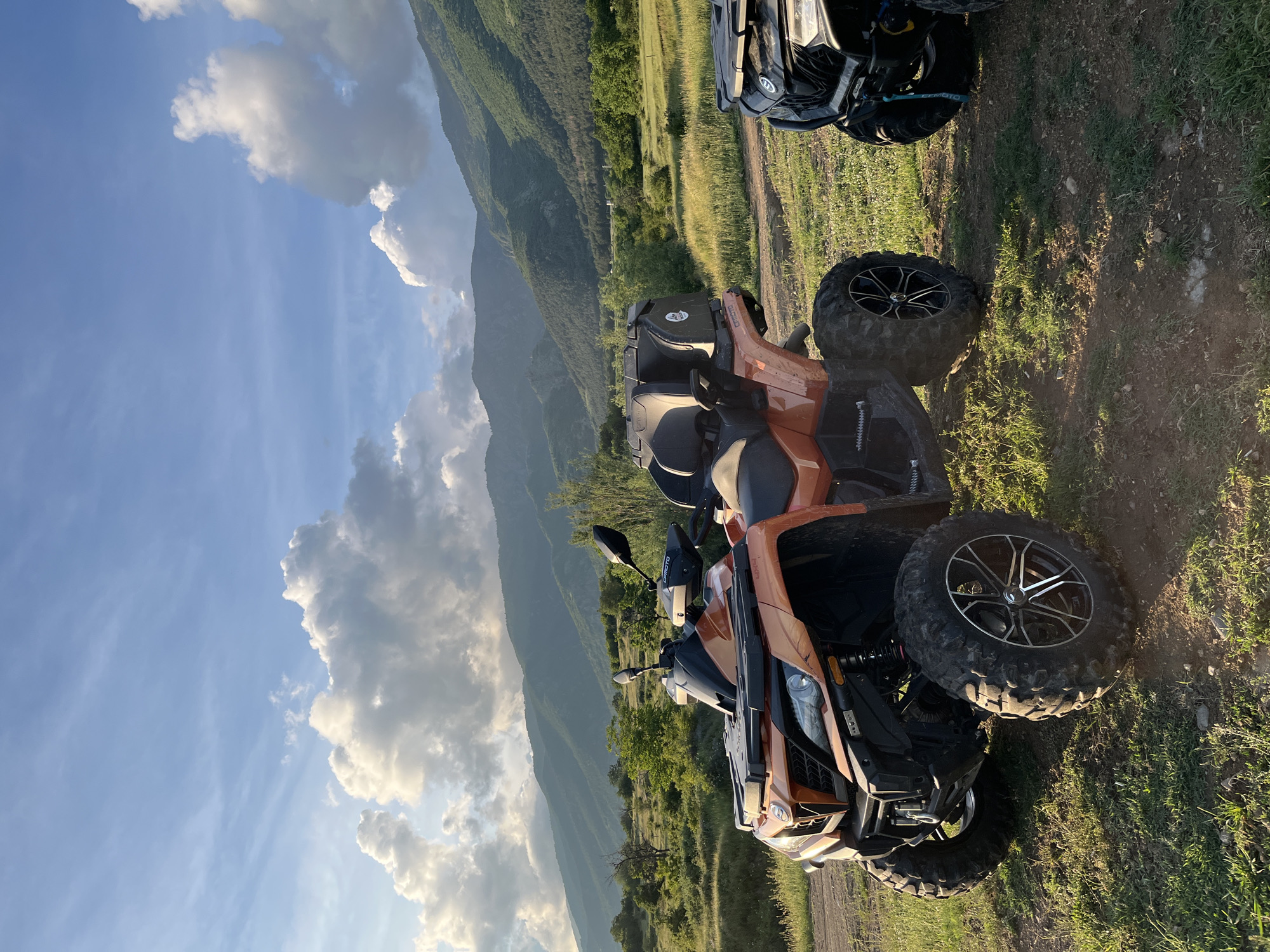 The image, originally rotated 90 degrees clockwise, captures two all-terrain vehicles (ATVs) parked on a dirt track in a mountainous rural landscape. The prominent ATV in the foreground is orange-brown, featuring large, knobby tires with distinctive black rims and silver spokes, a black seat, handlebars with mirrors, and visible headlights. A second, darker ATV (likely black) is partially visible behind it to the right.
The scene unfolds under a broad blue sky dotted with scattered white and grey cumulus clouds. Rolling green hills and mountains, densely covered with vegetation including grass and trees, dominate the background. The immediate ground consists of a rough dirt path interspersed with patches of green grass.
The lighting suggests late afternoon or early evening, with warm, soft sunlight casting gentle illumination and creating some shadows on the ground. The weather appears clear and pleasant. No people are visible in the frame. A small white "CFMOTO" logo is discernible on the front left side of the orange ATV, near the handlebar. The image depicts a tranquil moment during what appears to be an off-road adventure in a picturesque, verdant, mountainous region, consistent with the location of Anevo, Bulgaria.